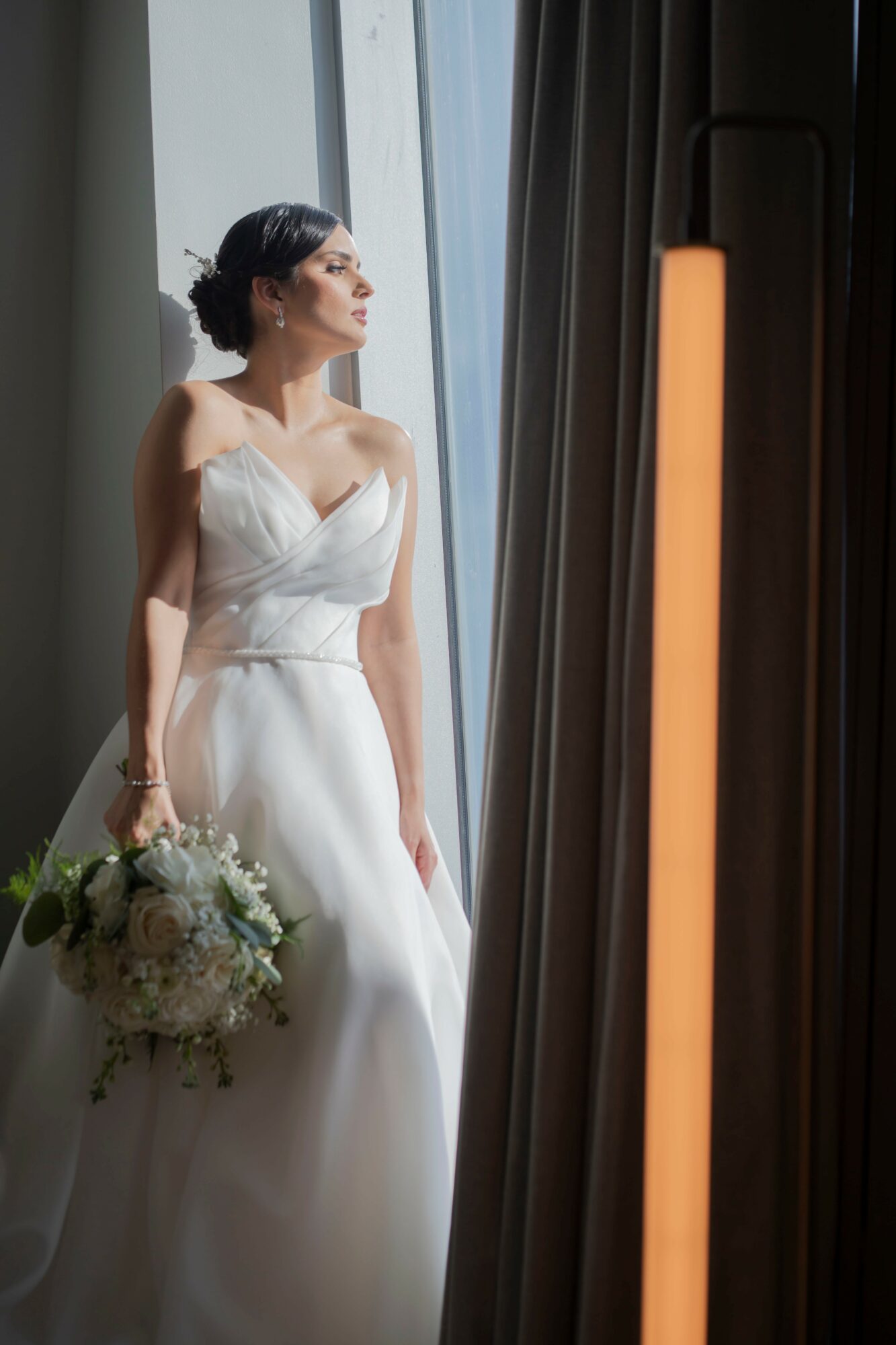 Woman in wedding dress holding bouquet, looking out window, sunlight illuminating her face, curtains partially drawn, indoor setting