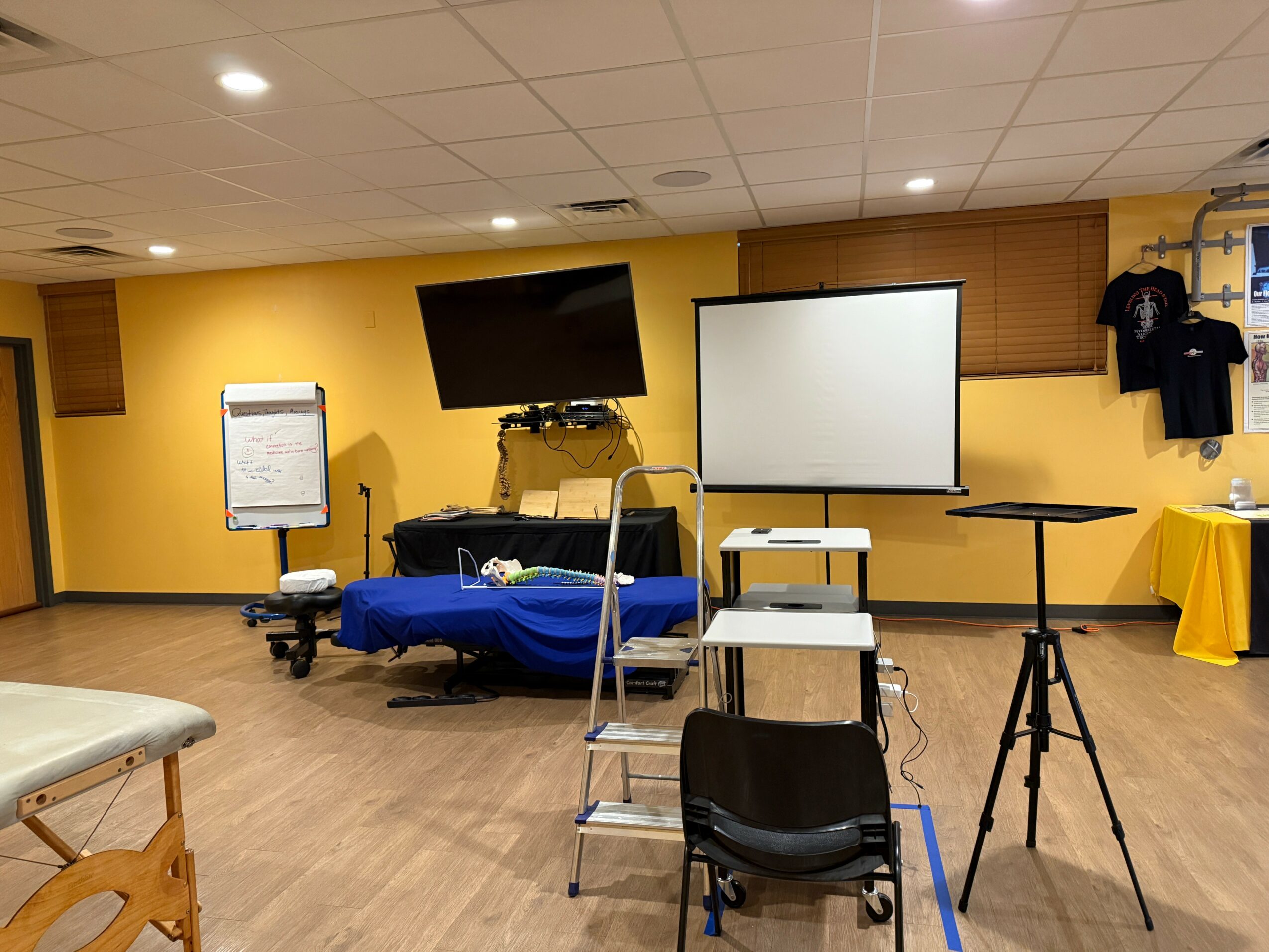 Room with yellow walls, TV, whiteboard, projector screen, and various equipment, set up for a presentation or meeting.
