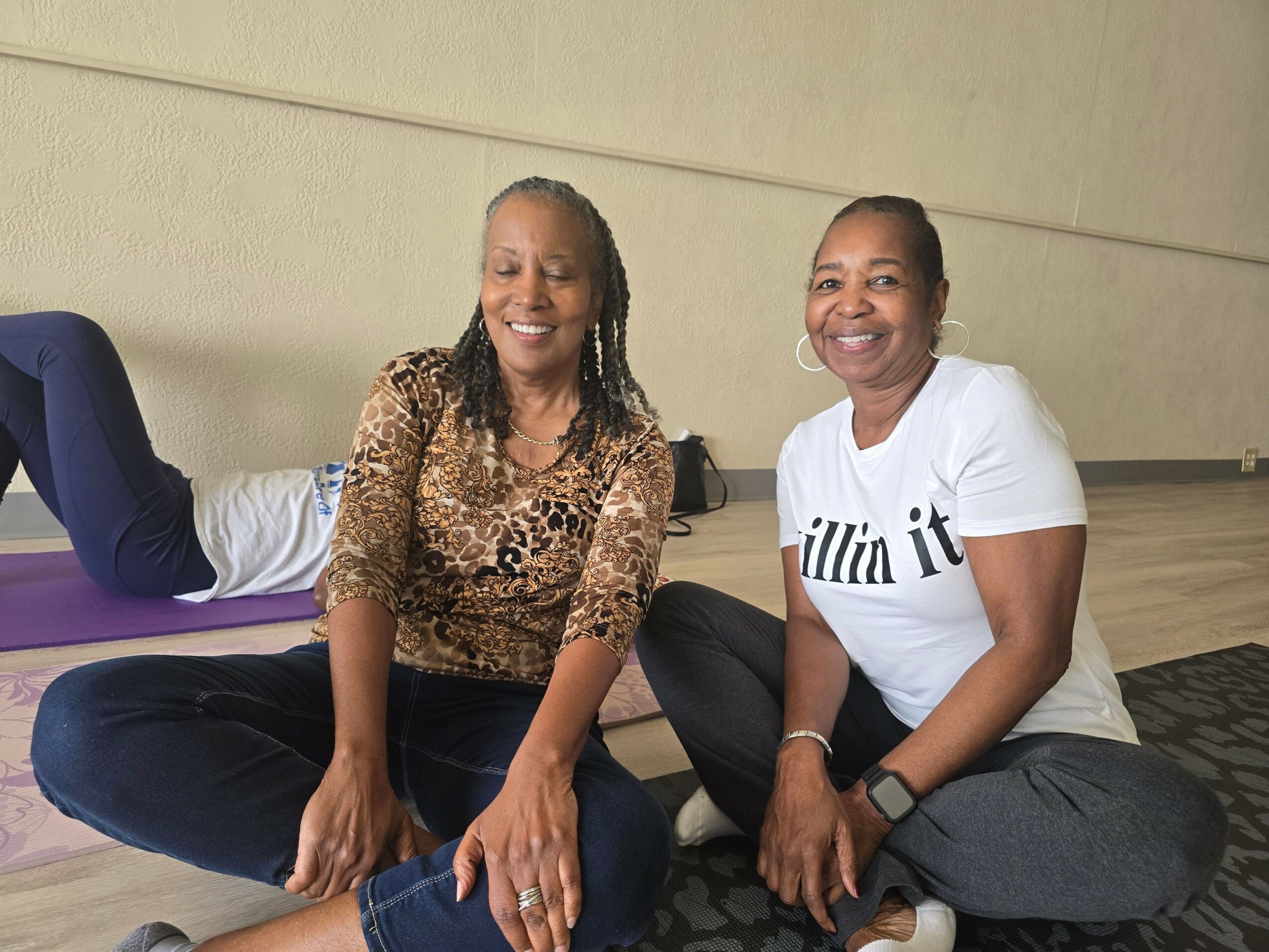 Two women sitting on a bench, smiling, with one wearing a white shirt and the other a leopard print top.