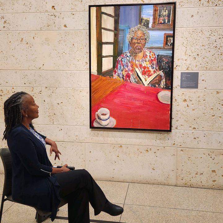 Woman sitting and observing a painting of an elderly woman reading a book, displayed on a wall in a gallery.