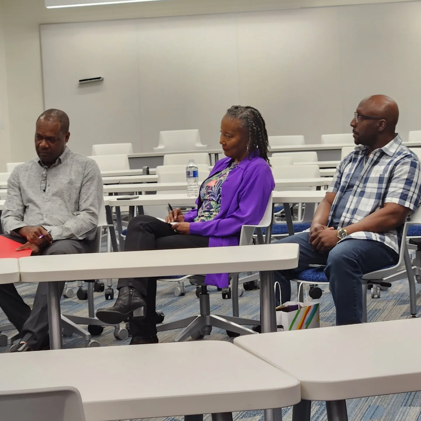 Three people sitting at a table in a classroom or conference room, with empty chairs and tables in the background.