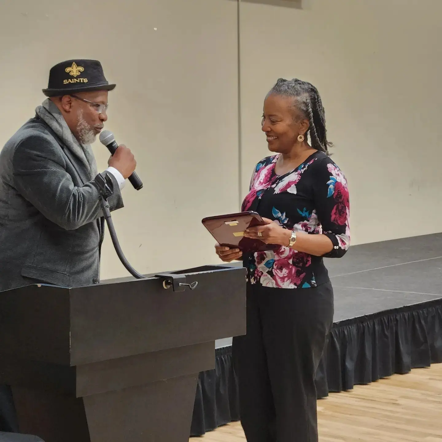 A man and woman stand on a stage, exchanging a smile. The man holds a microphone, and the woman holds a tablet.