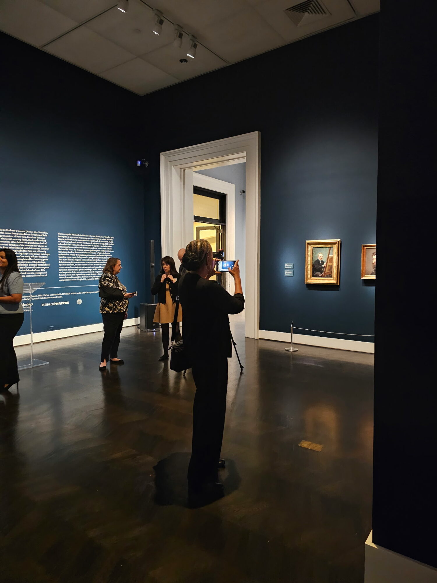 People viewing artwork in a dark gallery with blue walls and framed pictures, one person taking a photo.
