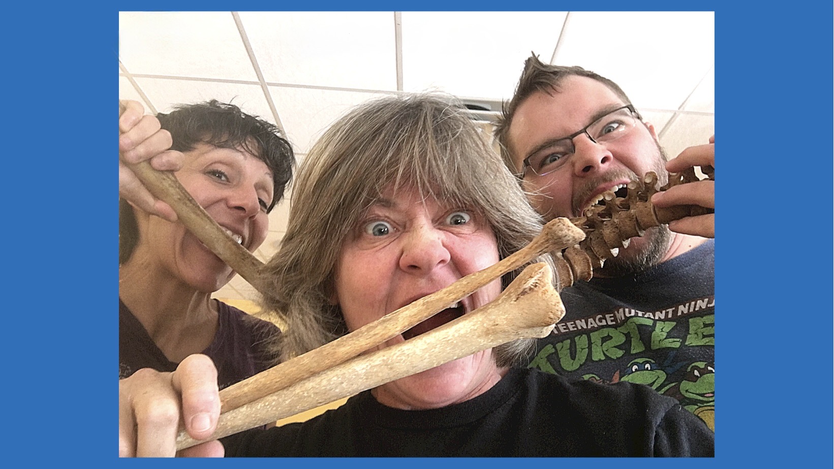 Three people with wide eyes biting into a large bone, smiling, in a room with a white ceiling and blue border.