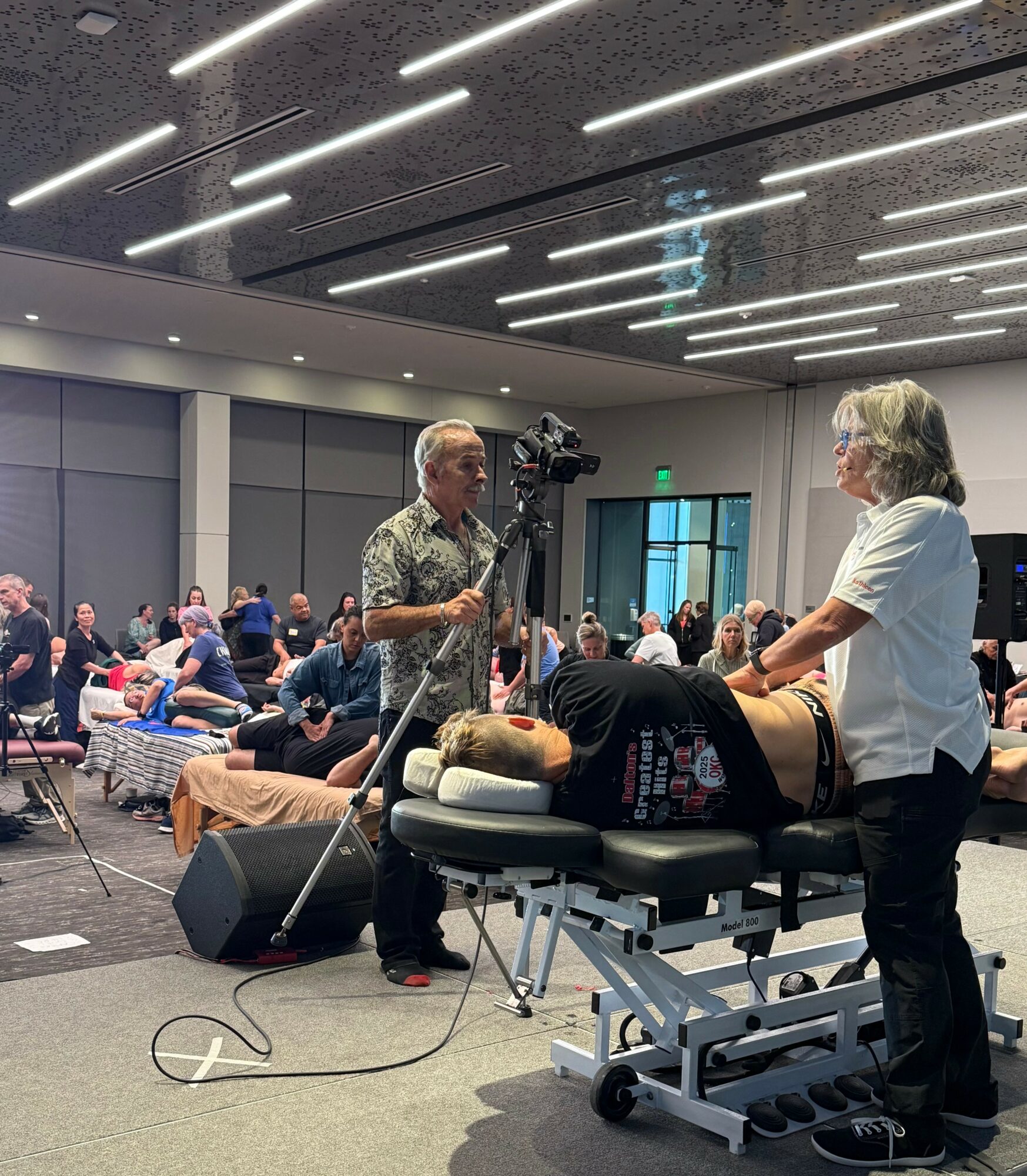 A person lying on a medical table with a pillow, being examined by a healthcare professional in a large room with many people seated. Fluorescent lights on the ceiling.