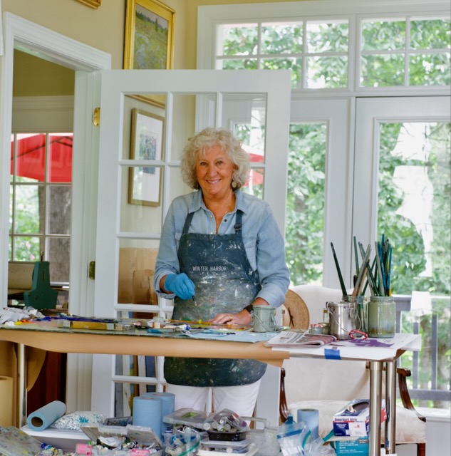 Older woman with curly white hair smiling, wearing apron and gloves, standing at a table with art supplies in a bright room.