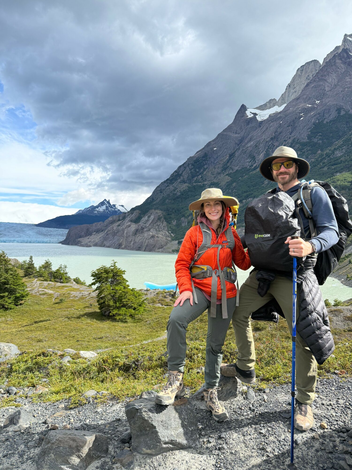Two hikers standing on rocks near a lake with mountains in the background, one in a black jacket and the other in an orange jacket.