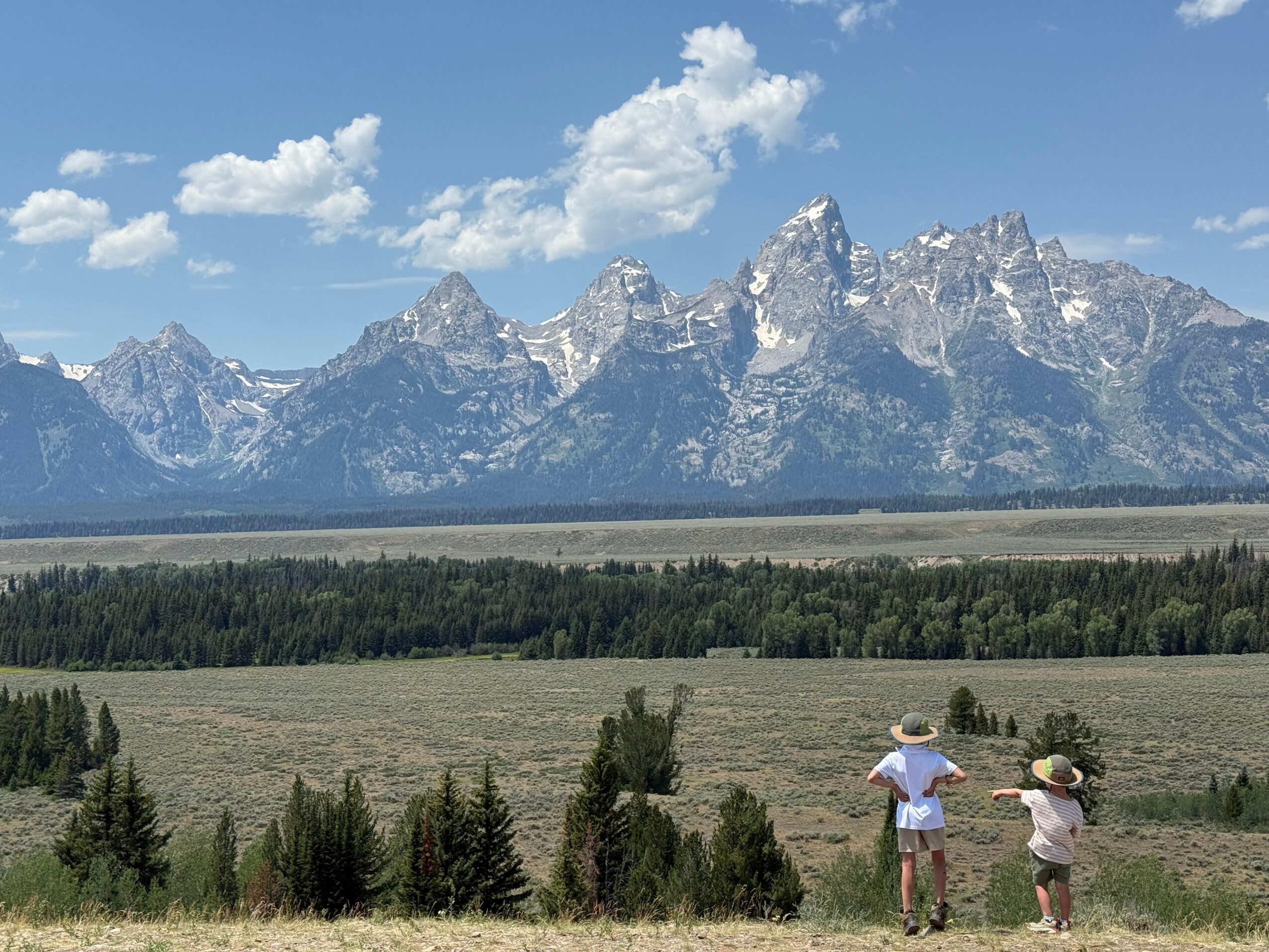 Two people wearing hats stand in a grassy field with mountains and a blue sky in the background.