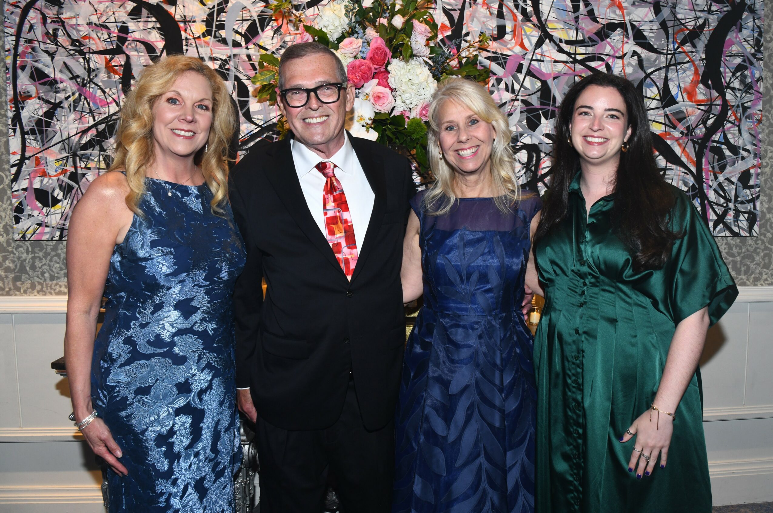 Four people standing together in front of a floral and abstract background, smiling, dressed in formal attire.