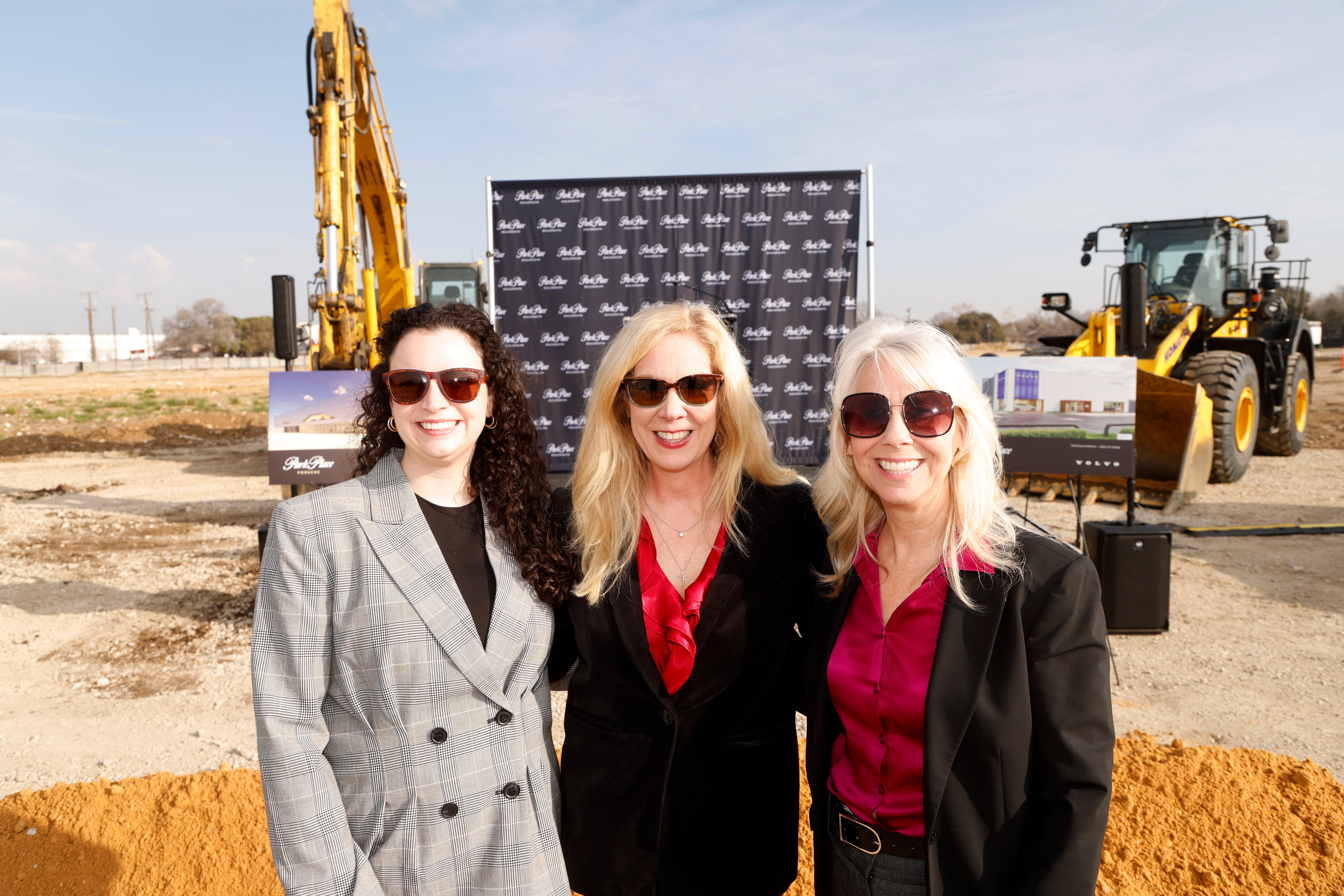 Three women with blonde and dark hair, wearing sunglasses and formal clothing, standing outdoors at a construction site.