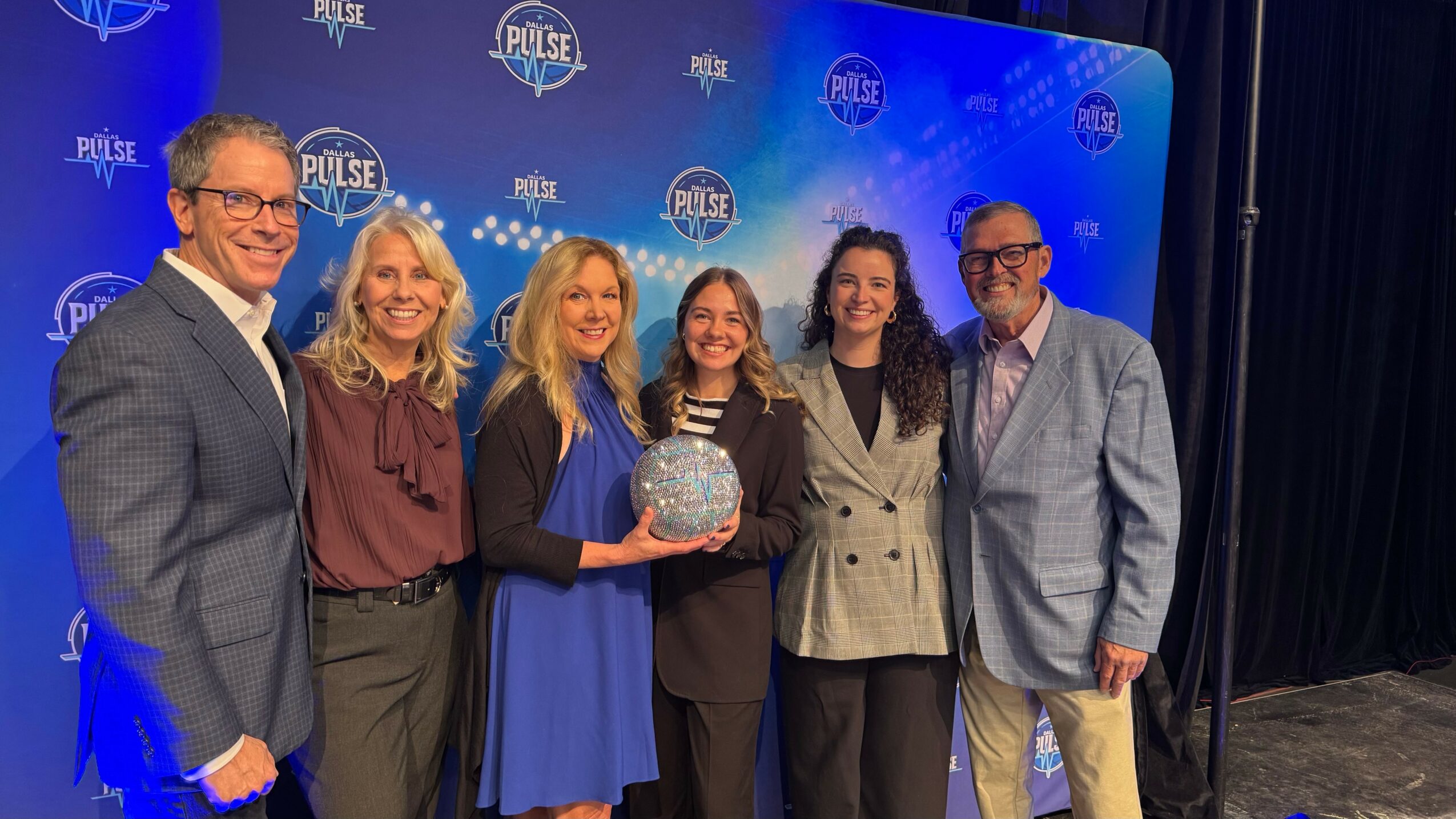 Group of six people standing on stage, smiling, with a blue backdrop and Pulse logo, one holding an award.