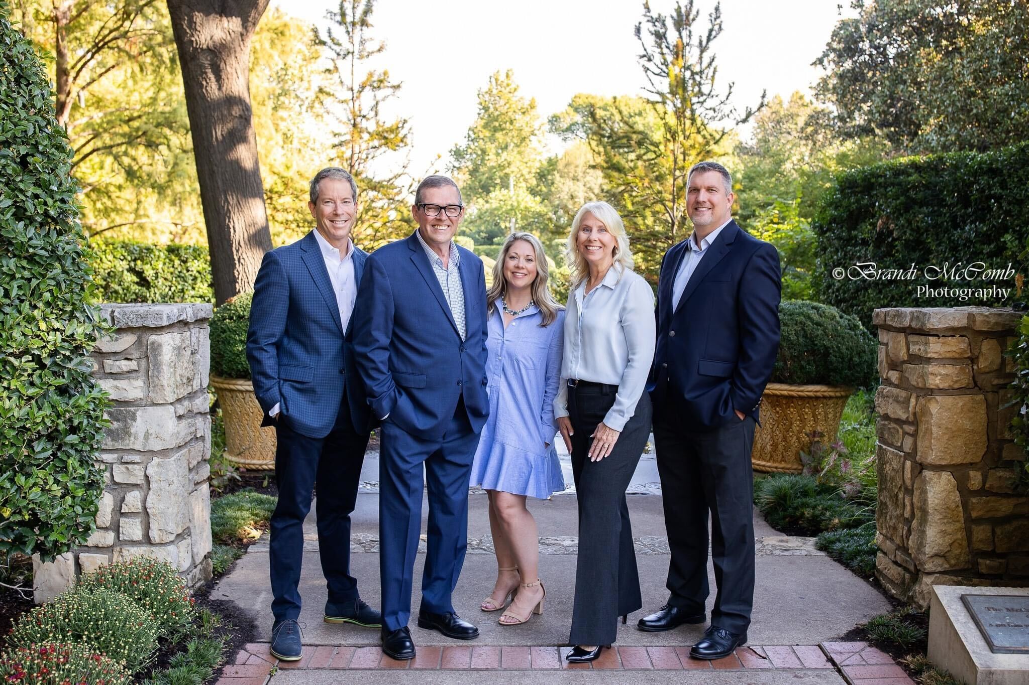 Four people standing outdoors on a paved path, surrounded by trees and greenery, smiling at the camera.