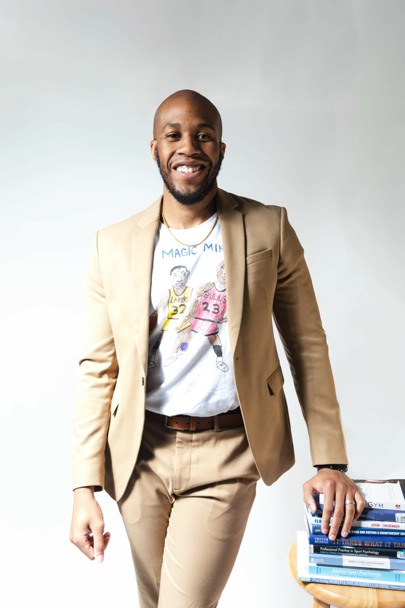 Smiling man in beige suit and graphic t-shirt standing against a plain background, with books on a table nearby.