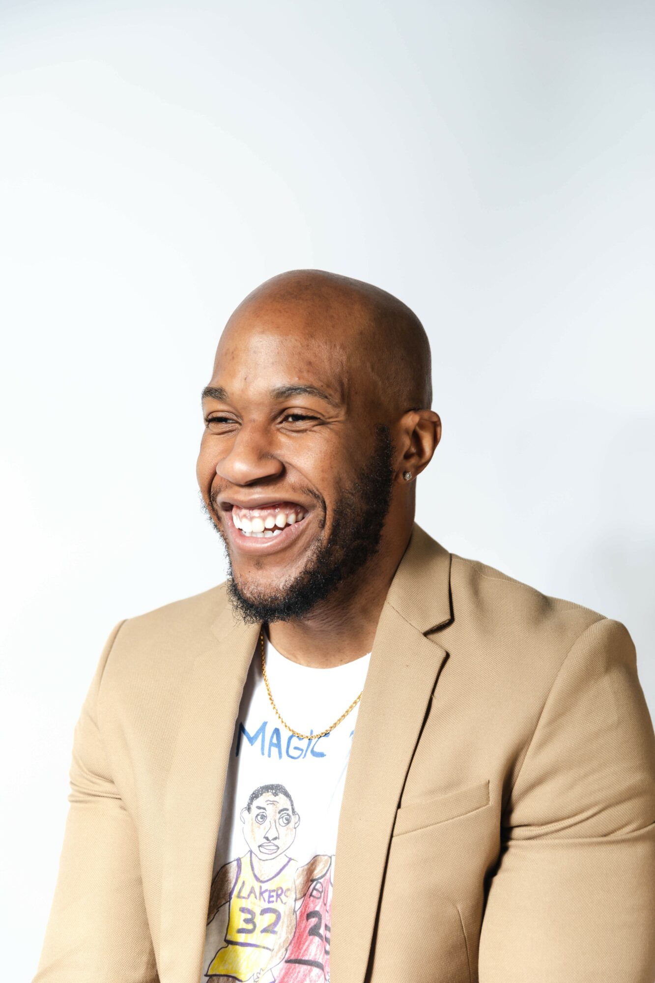 Smiling man with a beard and bald head wearing a beige blazer and graphic t-shirt, against a plain background.