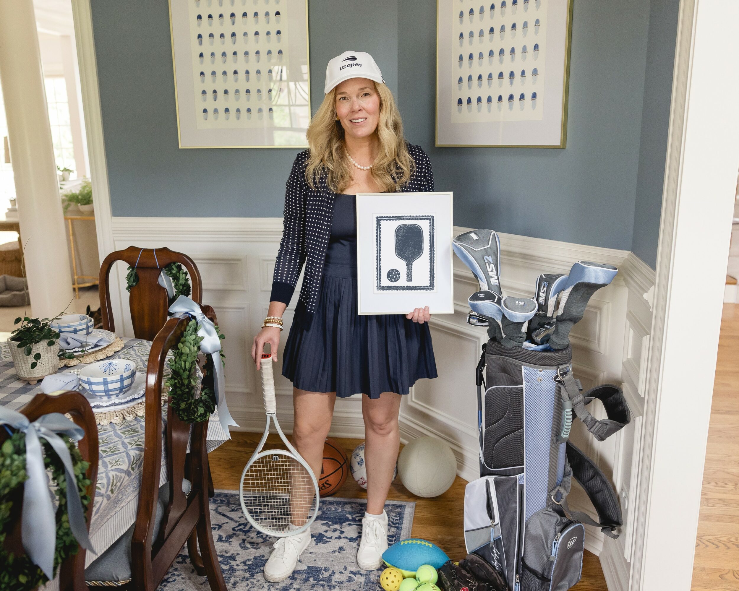 Woman in tennis outfit holding a framed headphone icon, standing in a dining room with golf clubs and tennis equipment.