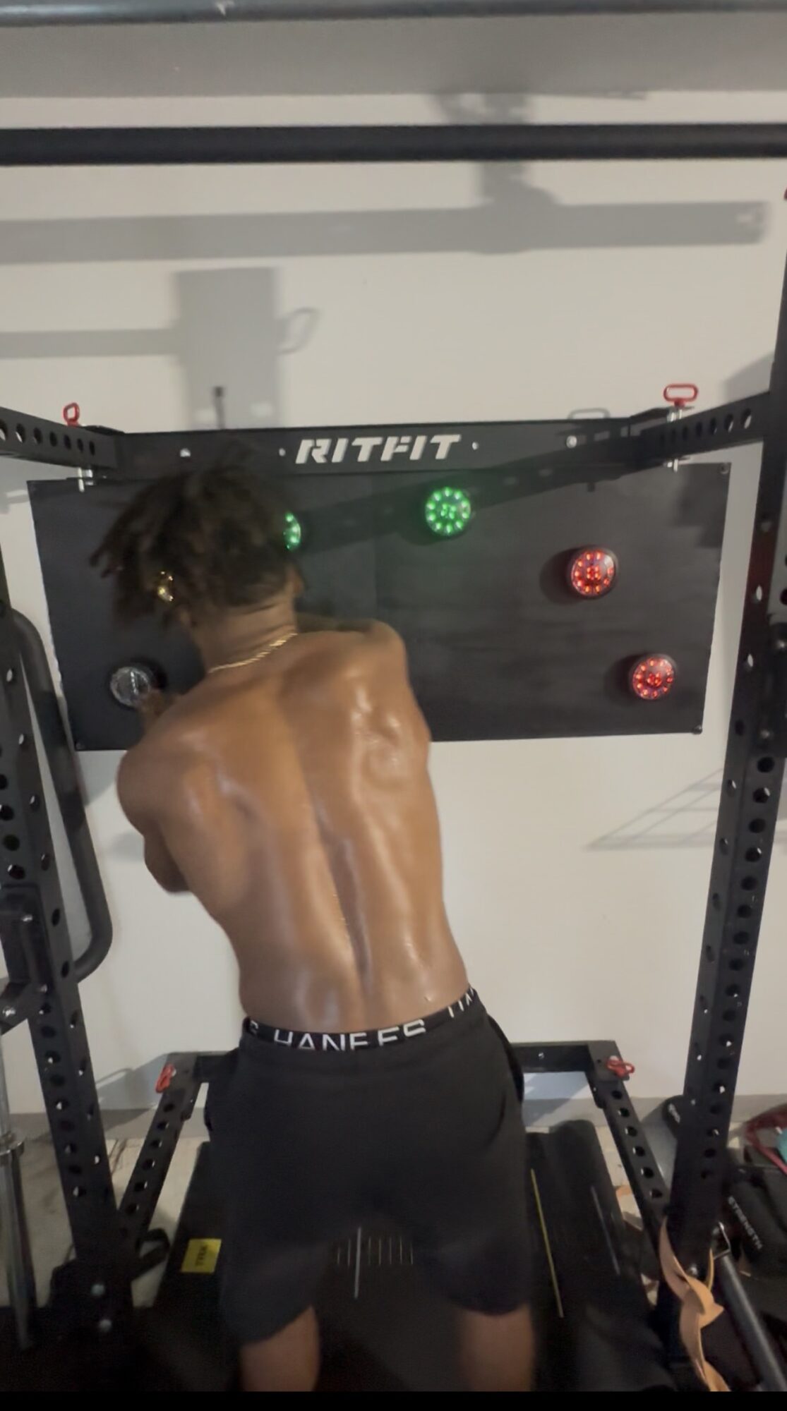 Person with back muscles visible, performing a workout on a black fitness machine in a gym. The person is shirtless and wearing black shorts.