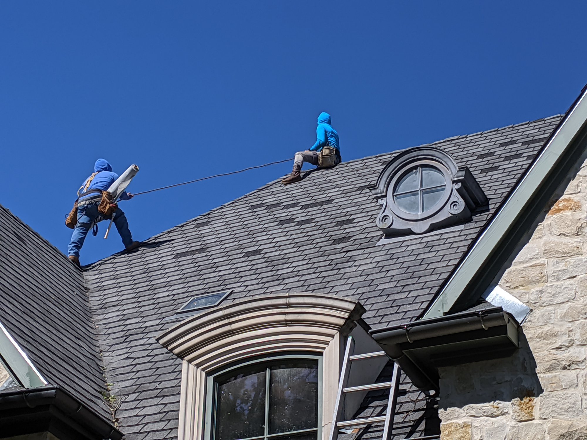 Two workers in blue clothing on a sloped roof, one holding a tool, with a circular window and chimney nearby.