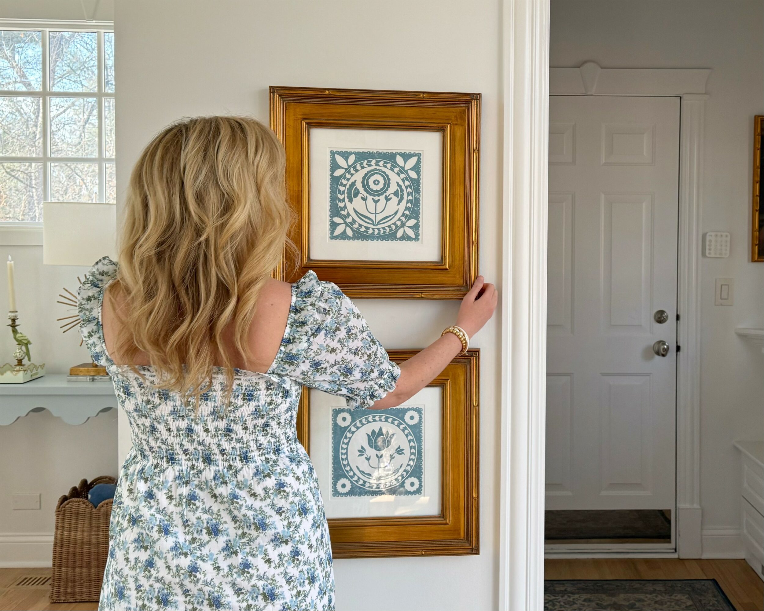Woman with wavy blonde hair looking at framed artwork on wall, in a bright room with a door nearby.