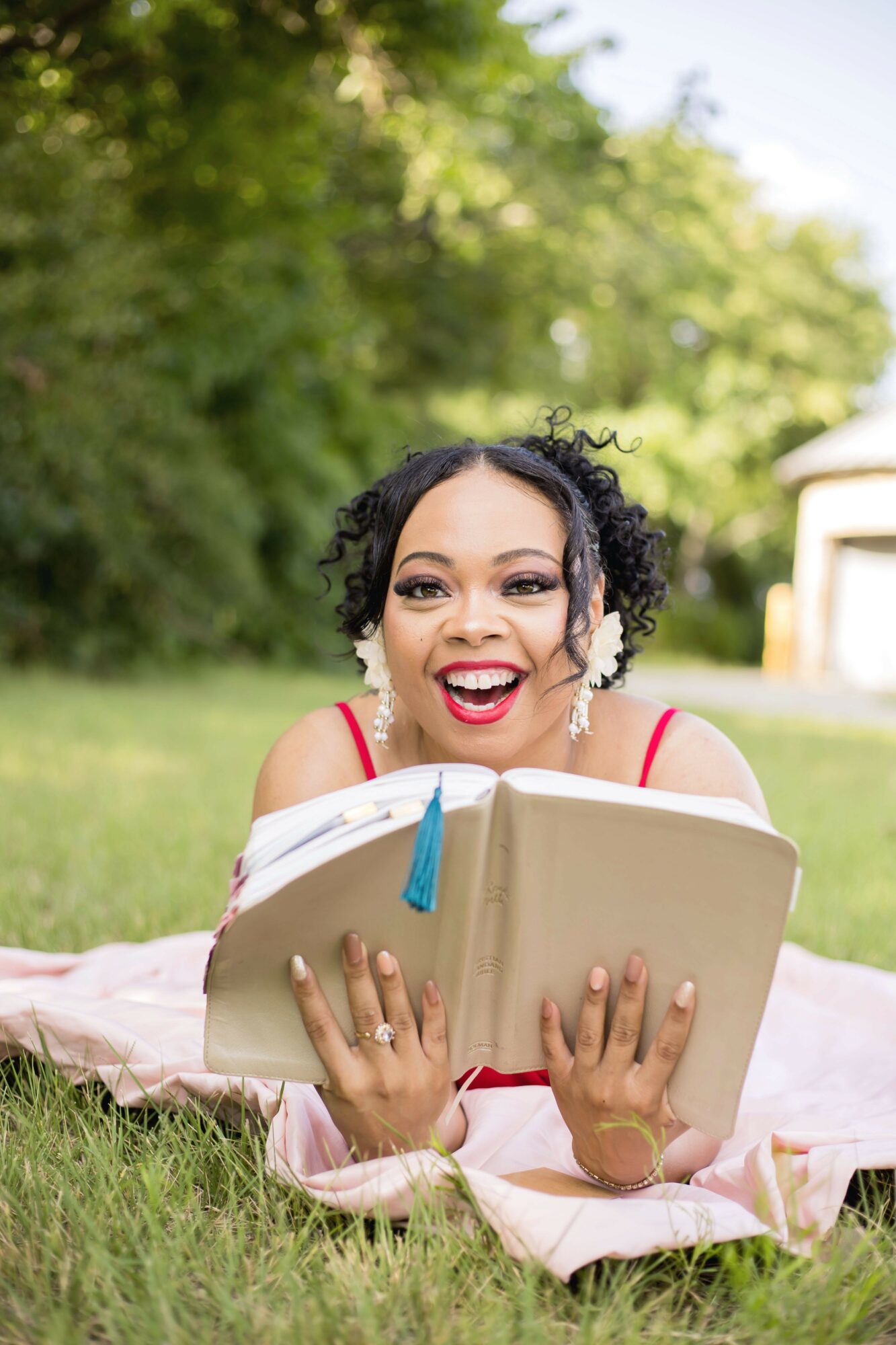 Woman lying on grass outdoors, holding a book, smiling, with trees and greenery in the background.