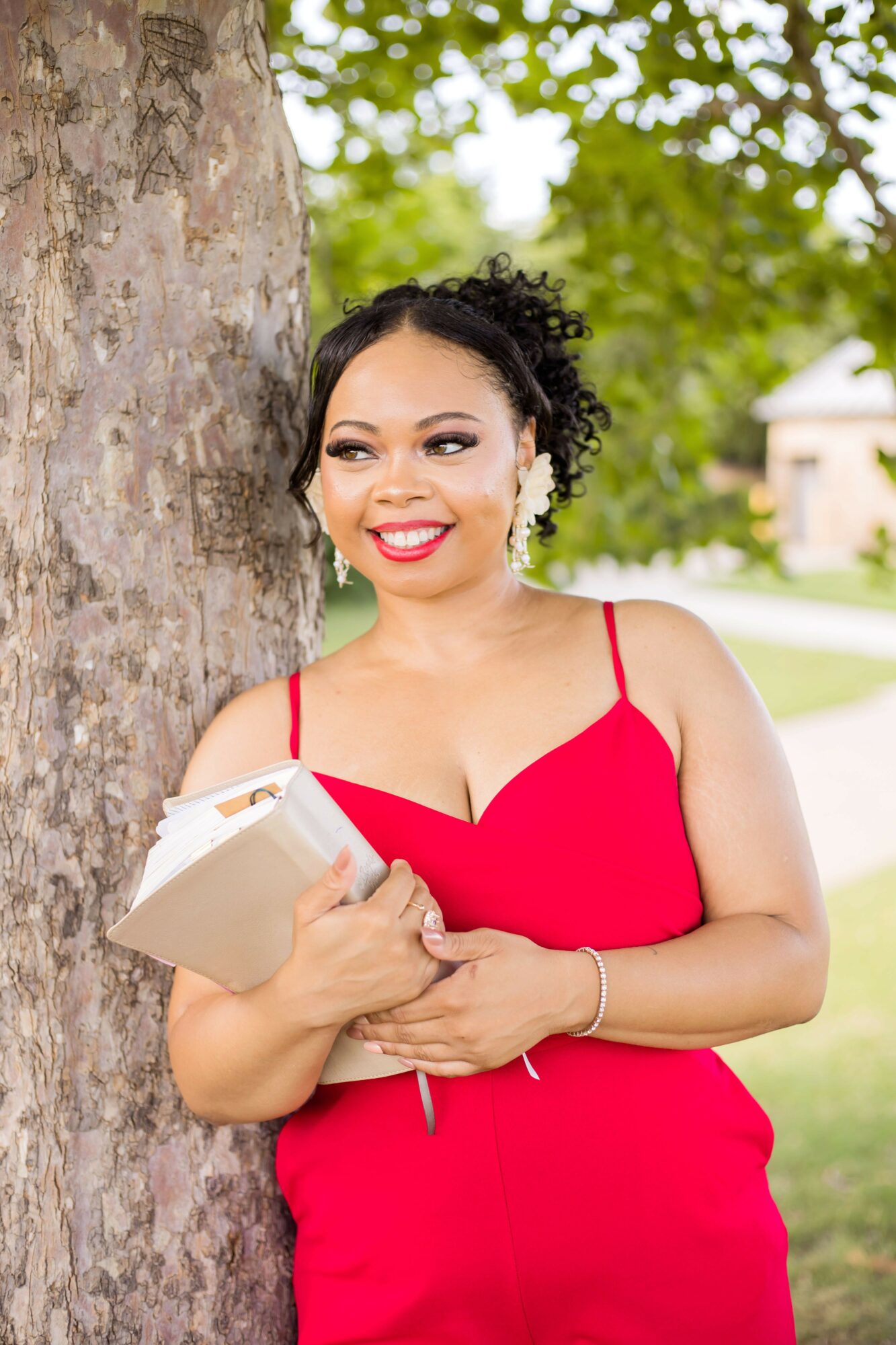 Woman in red dress holding a book outdoors, smiling, with trees and a stone wall in background.