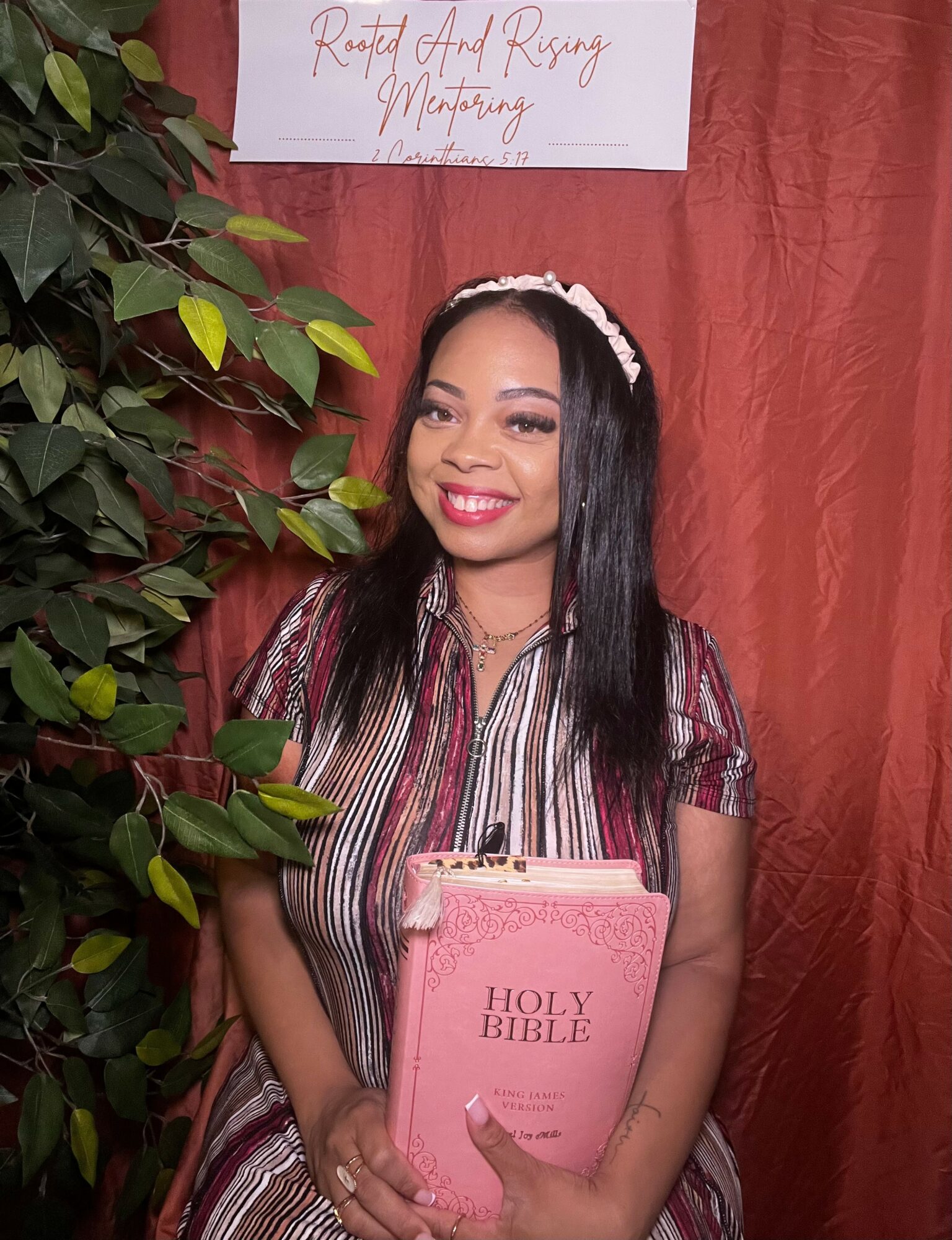 Smiling woman holding a pink Holy Bible, standing in front of a red curtain and greenery, wearing a striped dress and headband.