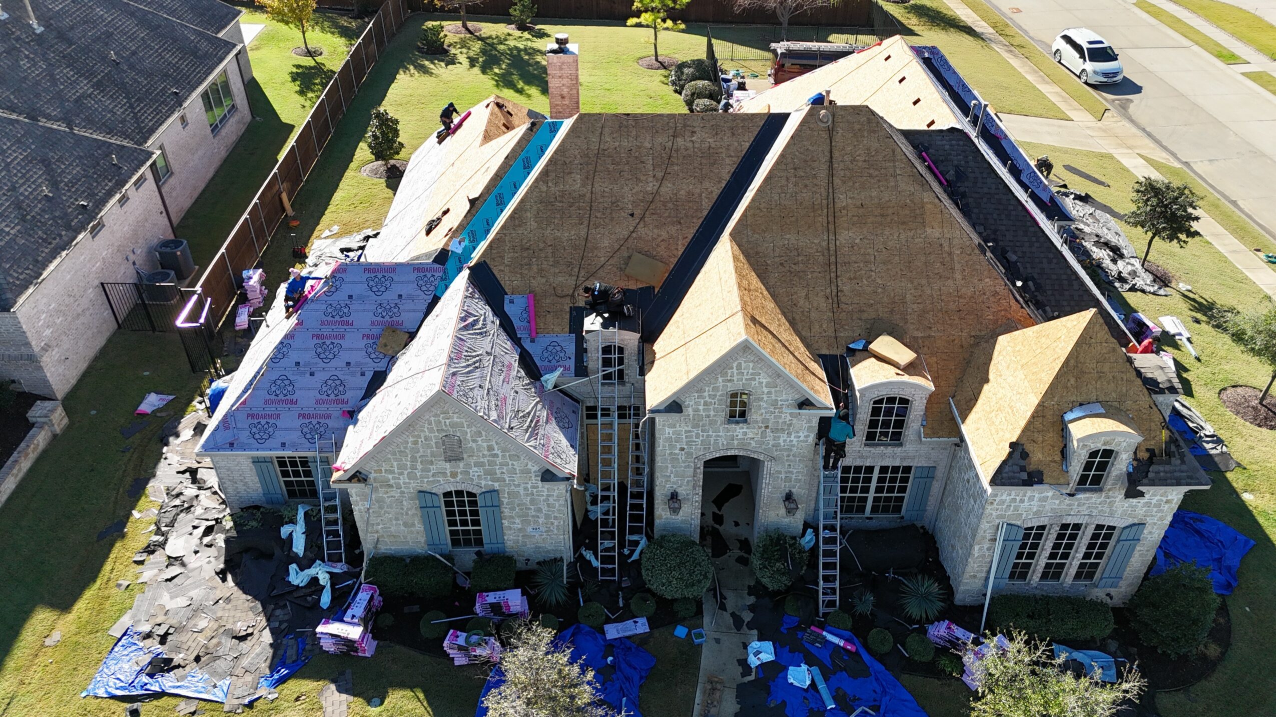 A large house under construction with scaffolding, surrounded by a lawn and neighboring houses, viewed from above.