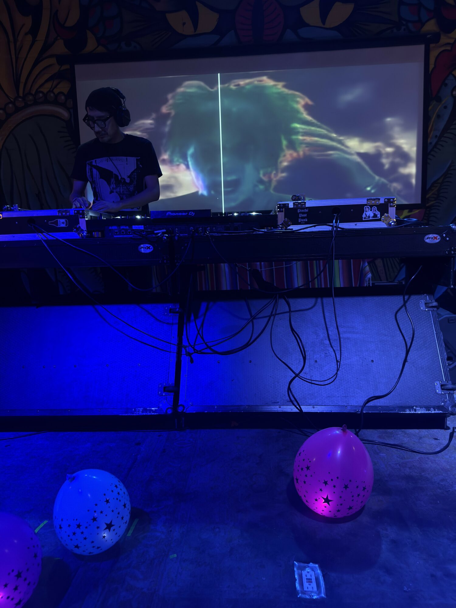Person DJing with a large screen displaying a colorful, abstract image behind them, and two balloons on the floor, one blue and one pink.