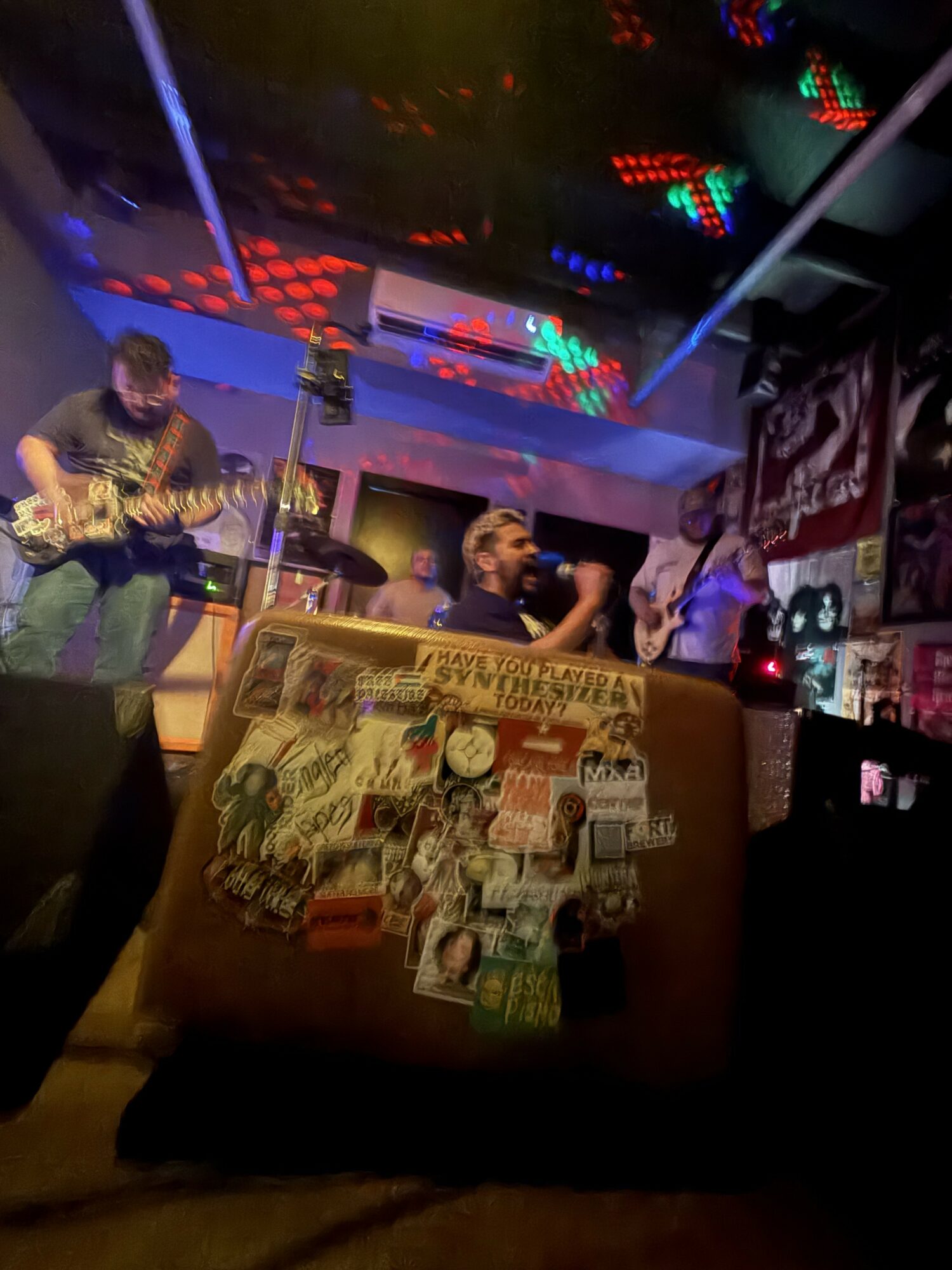 People playing guitars and singing on stage in a dimly lit venue with colorful lights and posters on the wall.