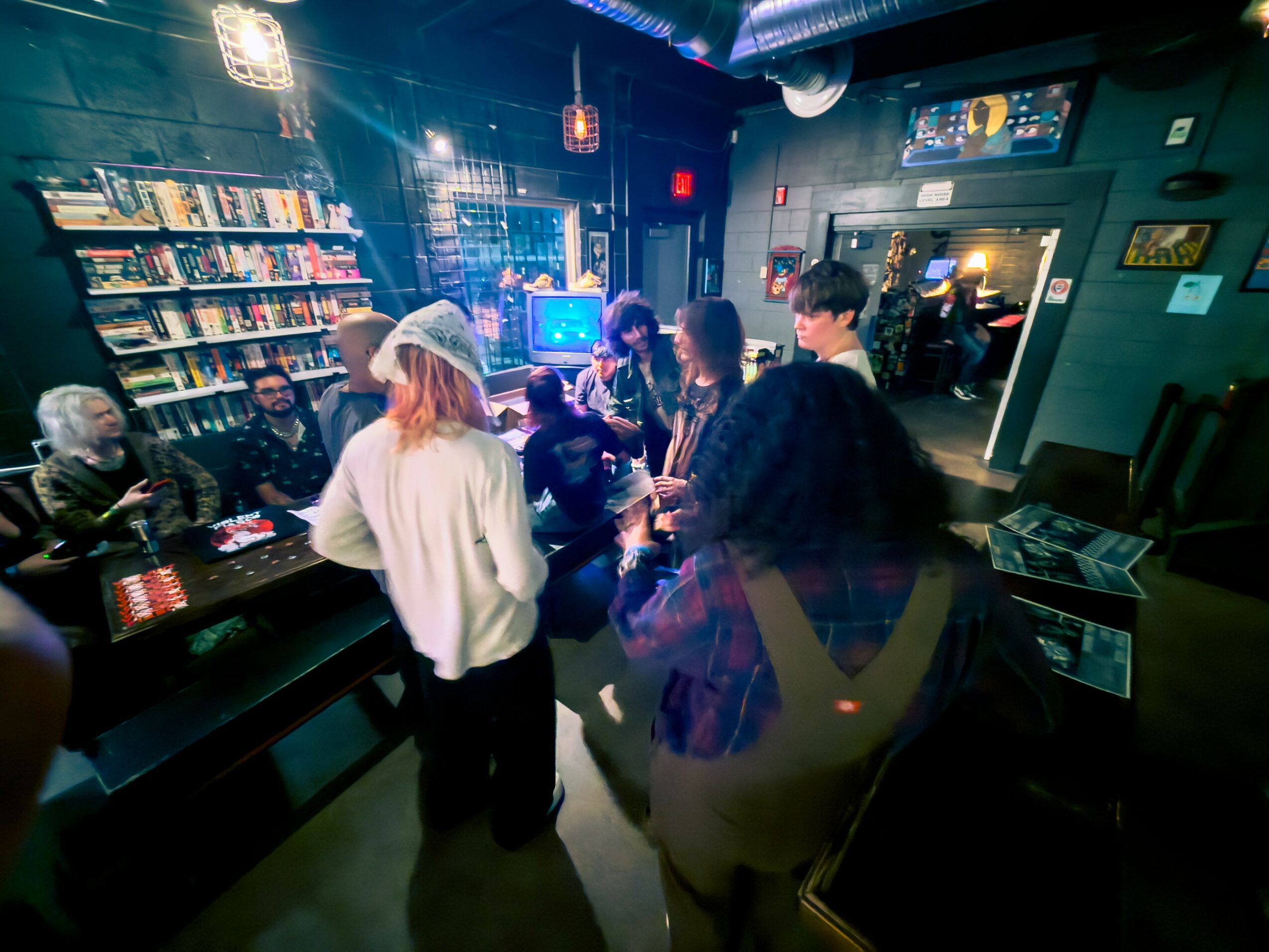 People gather around a bar in a dimly lit, modern space with bookshelves and a television screen, engaging in conversation.