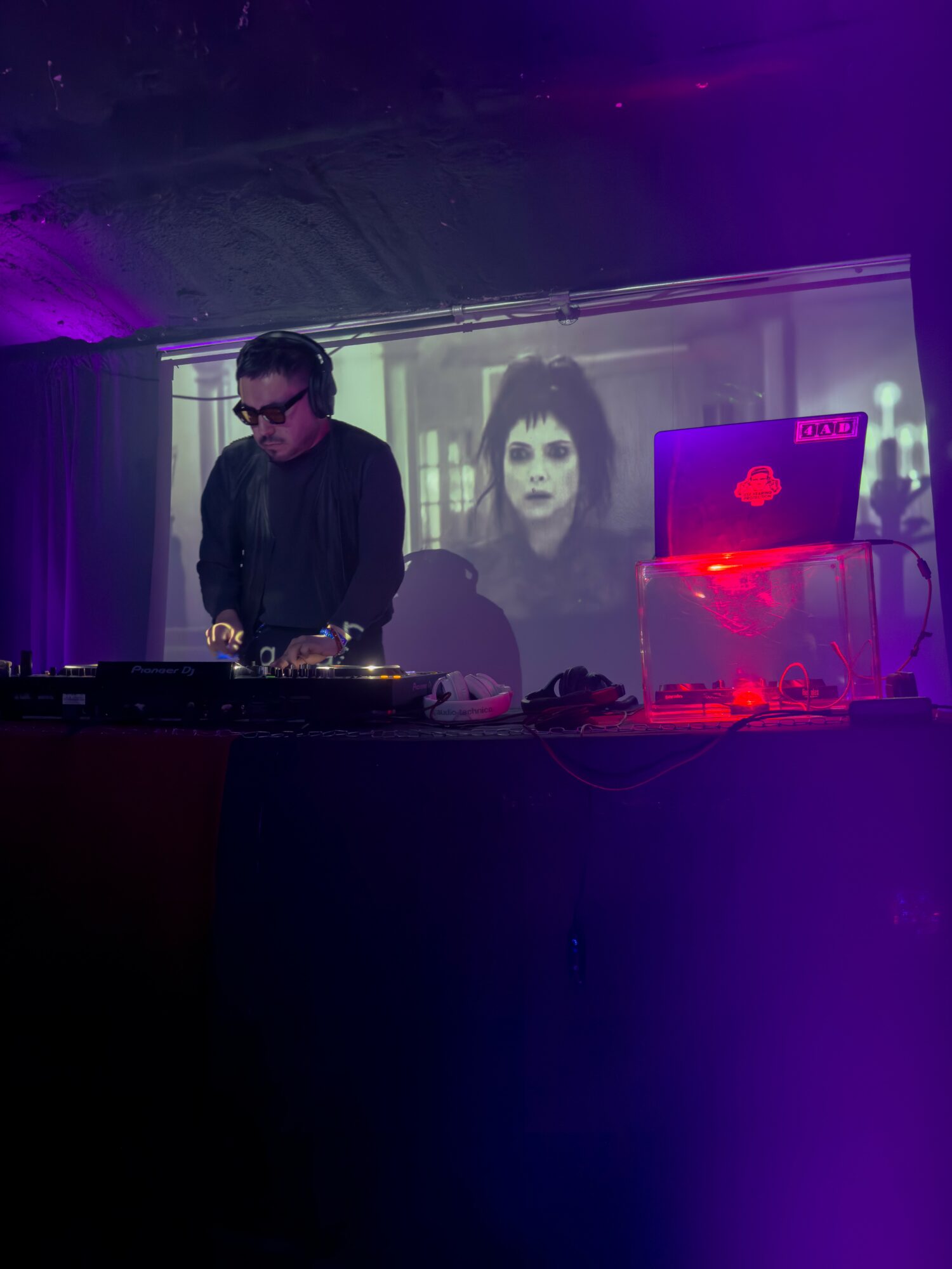 DJ with headphones at a booth, large screen behind showing a woman's face, purple and blue lighting, electronic equipment visible.