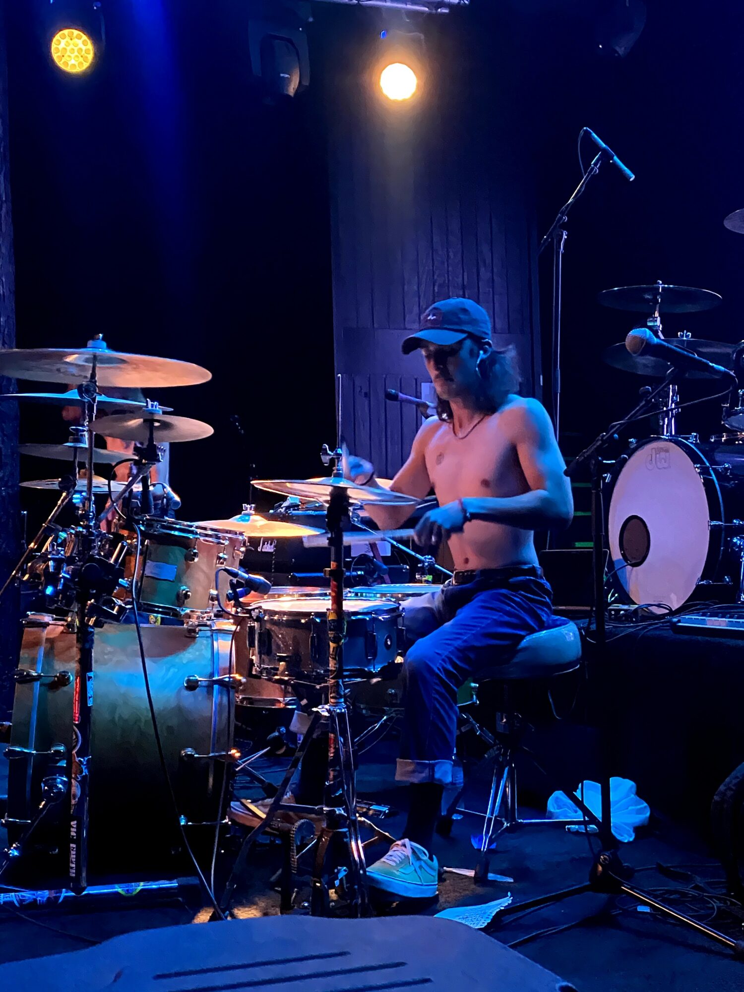 Female drummer playing drums on stage with blue and yellow lighting, wearing a cap and sleeveless top.