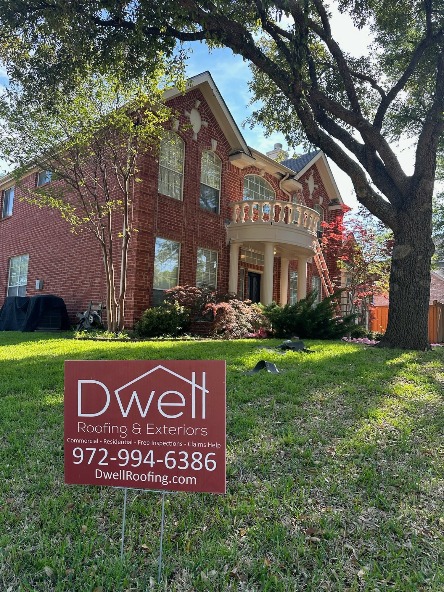 Two-story brick house with a front porch, surrounded by trees and a lawn, with a Dwell Roofing sign in the yard.
