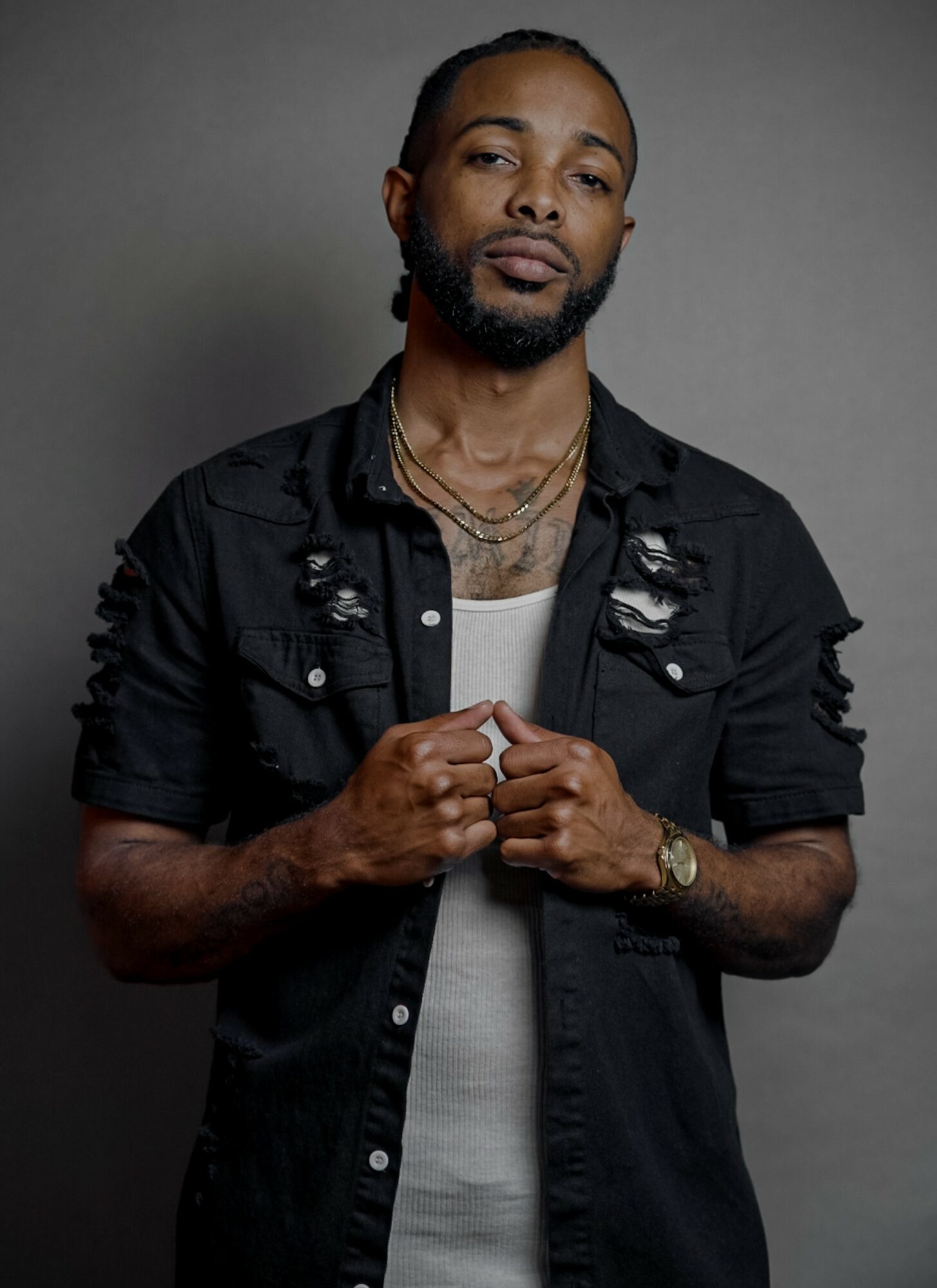Man with a beard and braided hair, wearing a black distressed shirt over a white top, standing against a gray background.