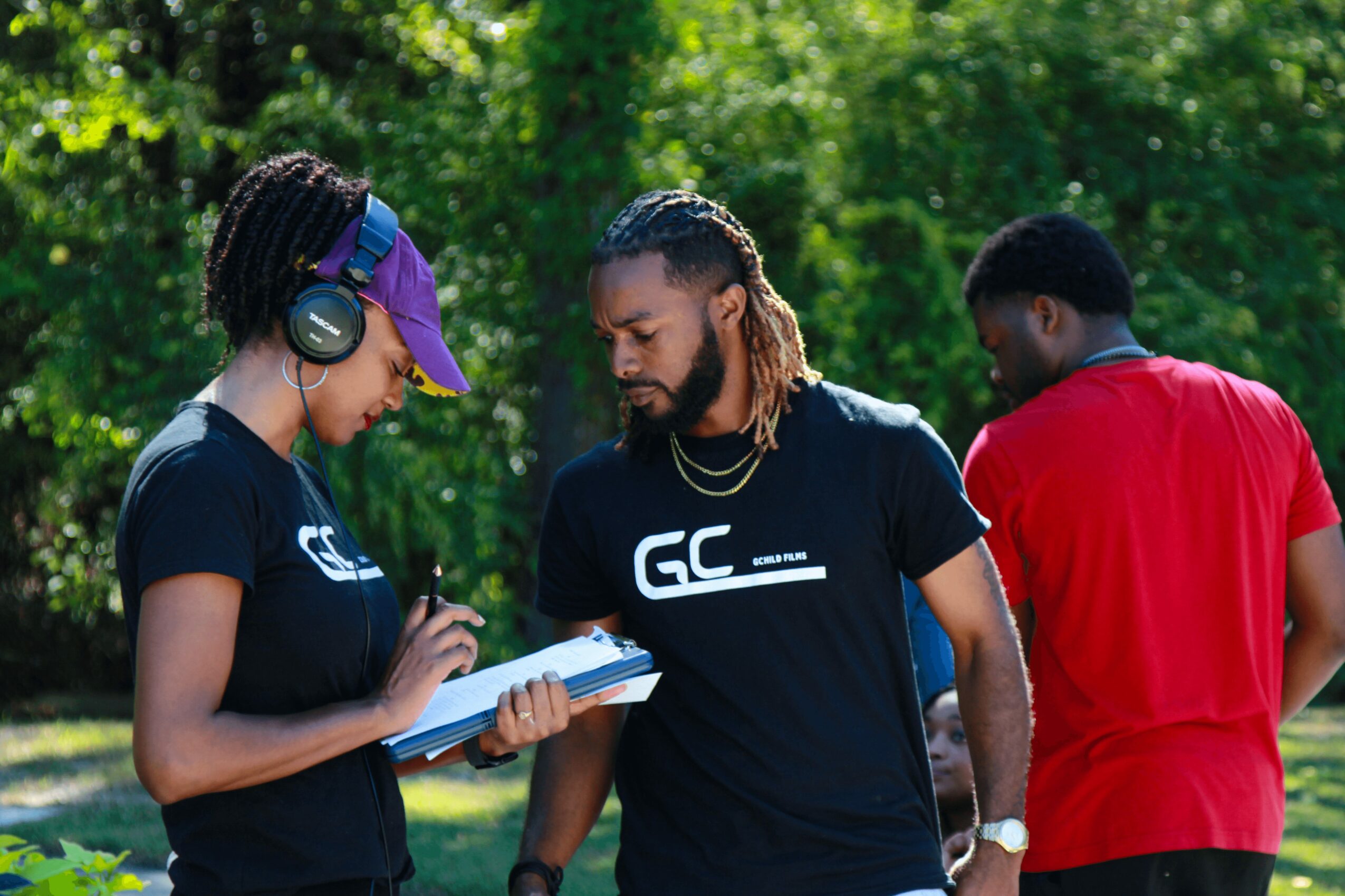 Three people outdoors, two wearing black shirts, one in red, with trees in the background.