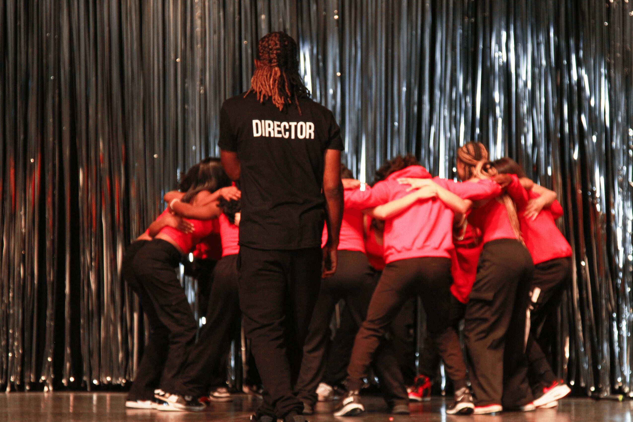Group of people in red and black clothing huddled together on stage with a person labeled 'Director' in front, silver curtain background.