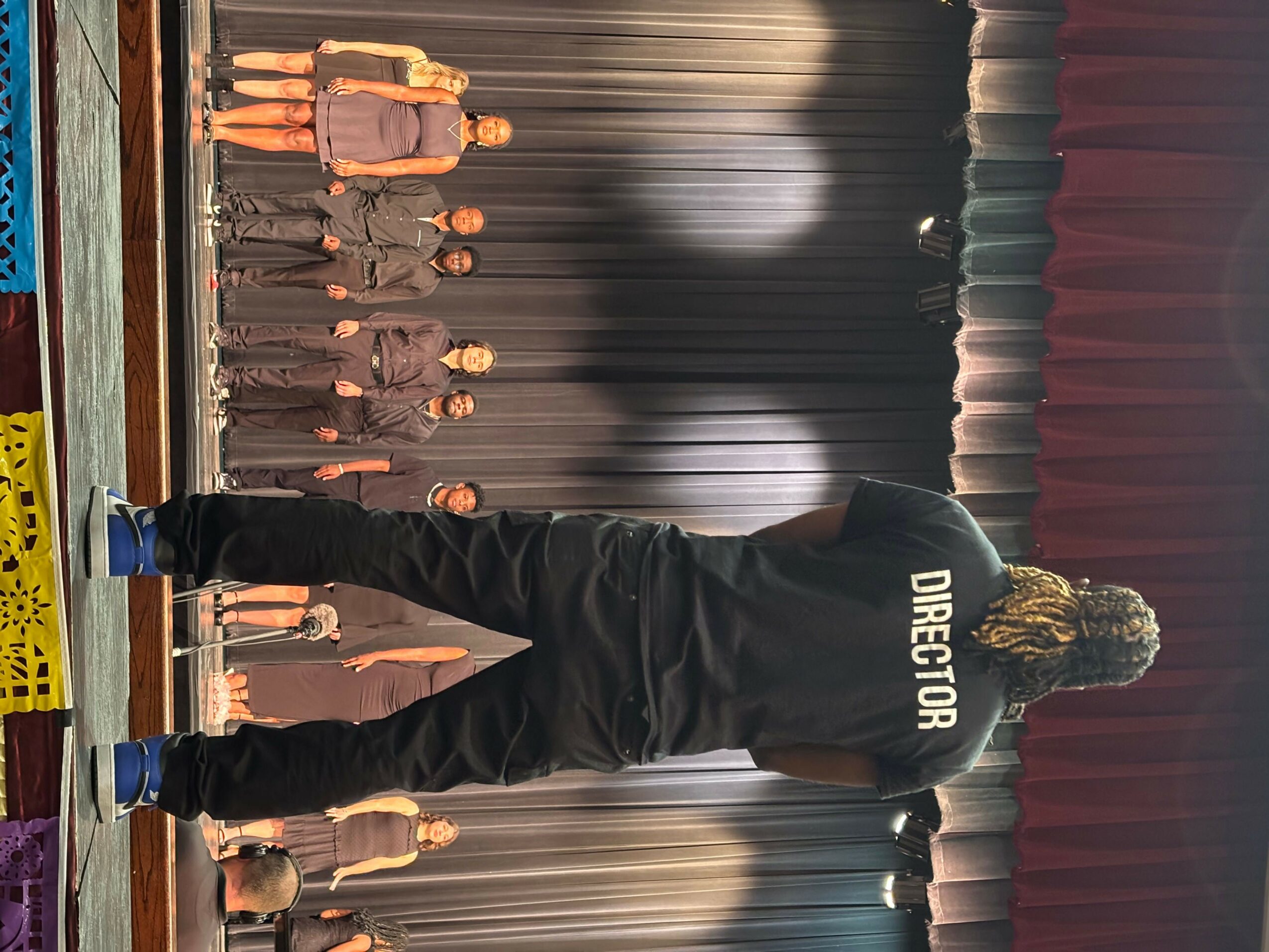Person with curly hair wearing a black hoodie labeled 'DIRECTOR' standing on stage facing an audience.