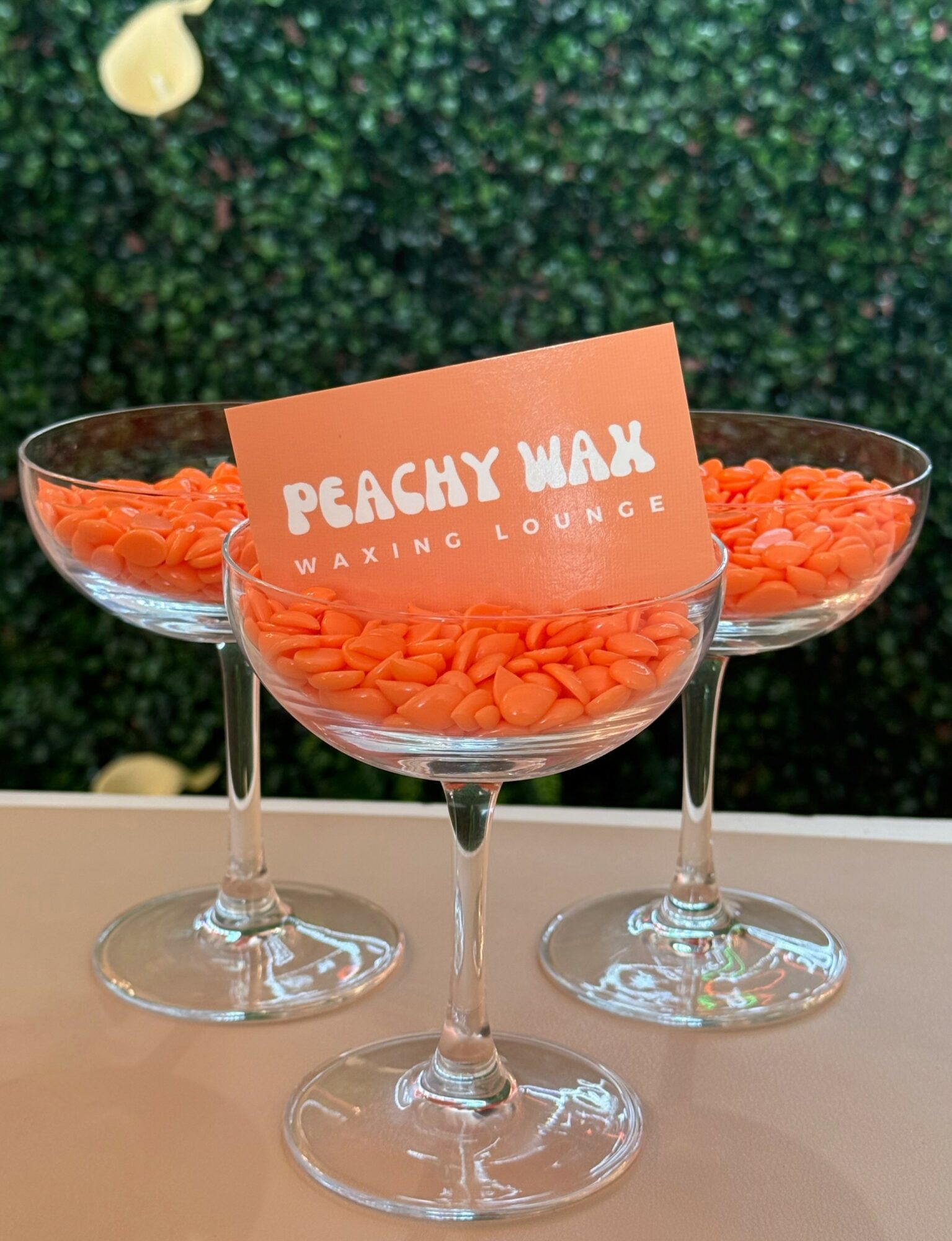 Three glass bowls filled with orange wax melts, with a peach-colored sign reading 'PEACHY WAX WAXING LOUNGE' in front, on a beige surface.