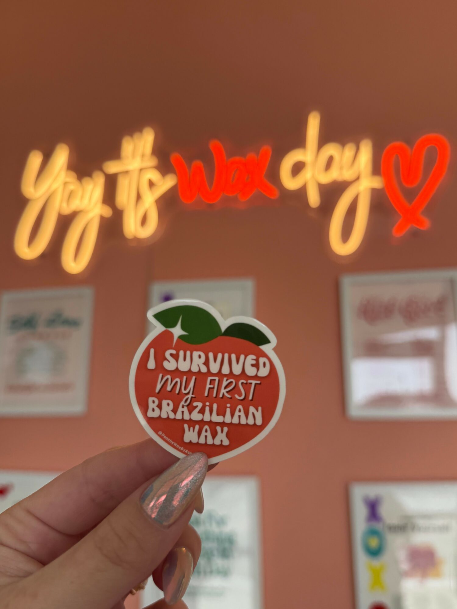 Hand holding a sticker with a red and white design, text about surviving a zombie apocalypse, in front of a pink wall with framed pictures