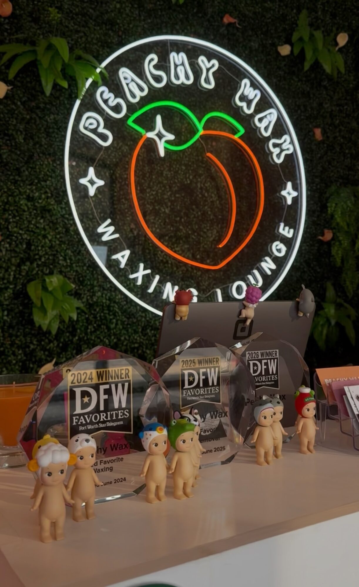 Neon sign with a peach and text, surrounded by small stars, above a table with plush toys and magazines.