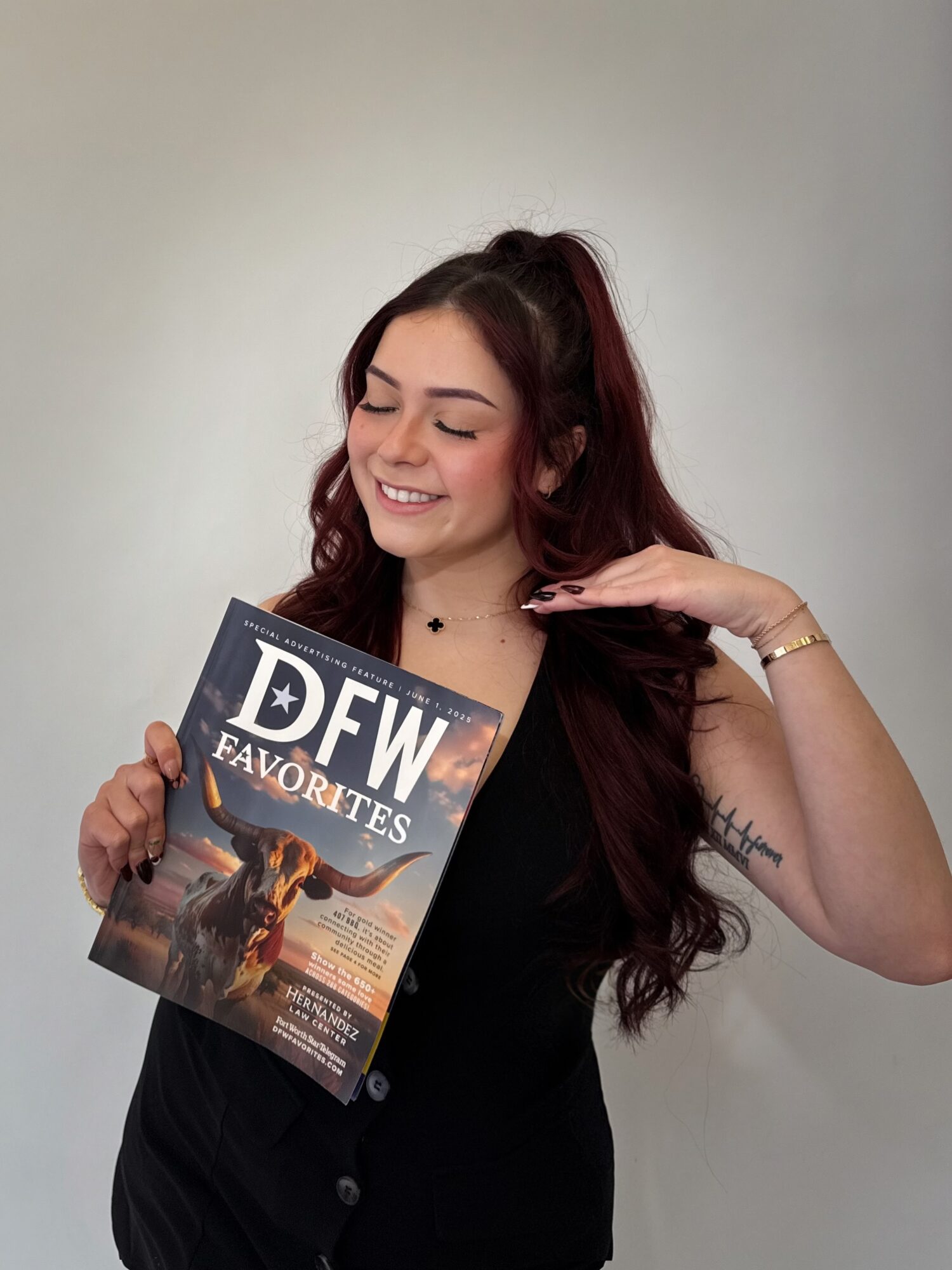 Young woman with long dark hair smiling, holding a magazine titled 'DFW FAVORITES', wearing a black top, against a plain background.