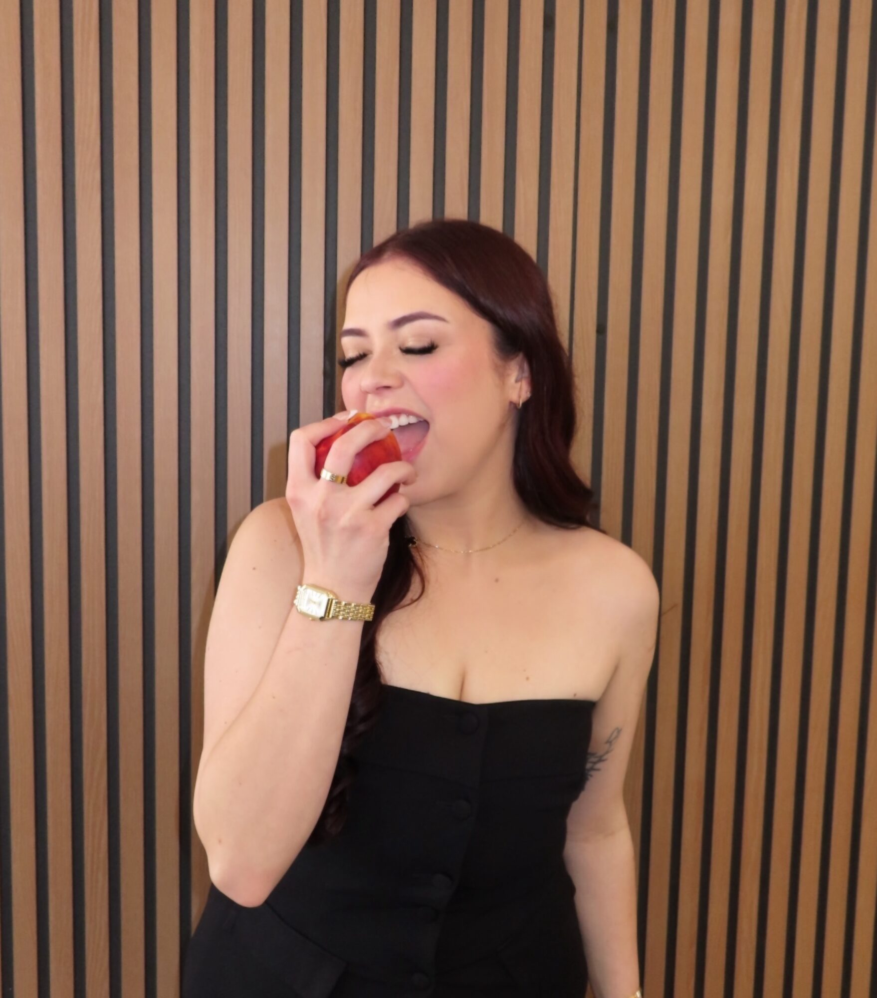 Woman with long dark hair in a strapless black dress bites into a red apple against a wooden striped background.