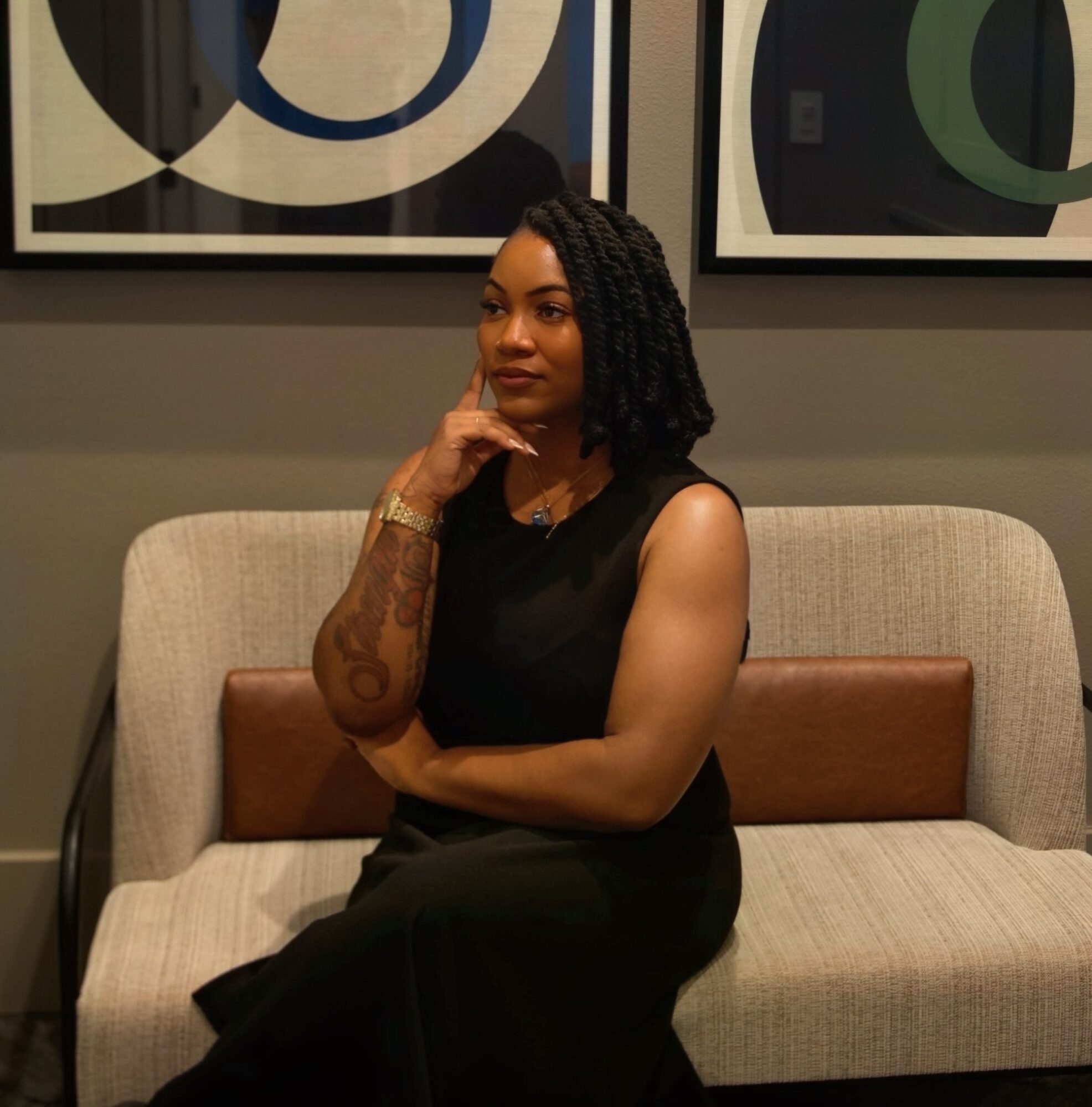Woman with curly hair sitting on a beige and brown sofa, with abstract artwork behind her.