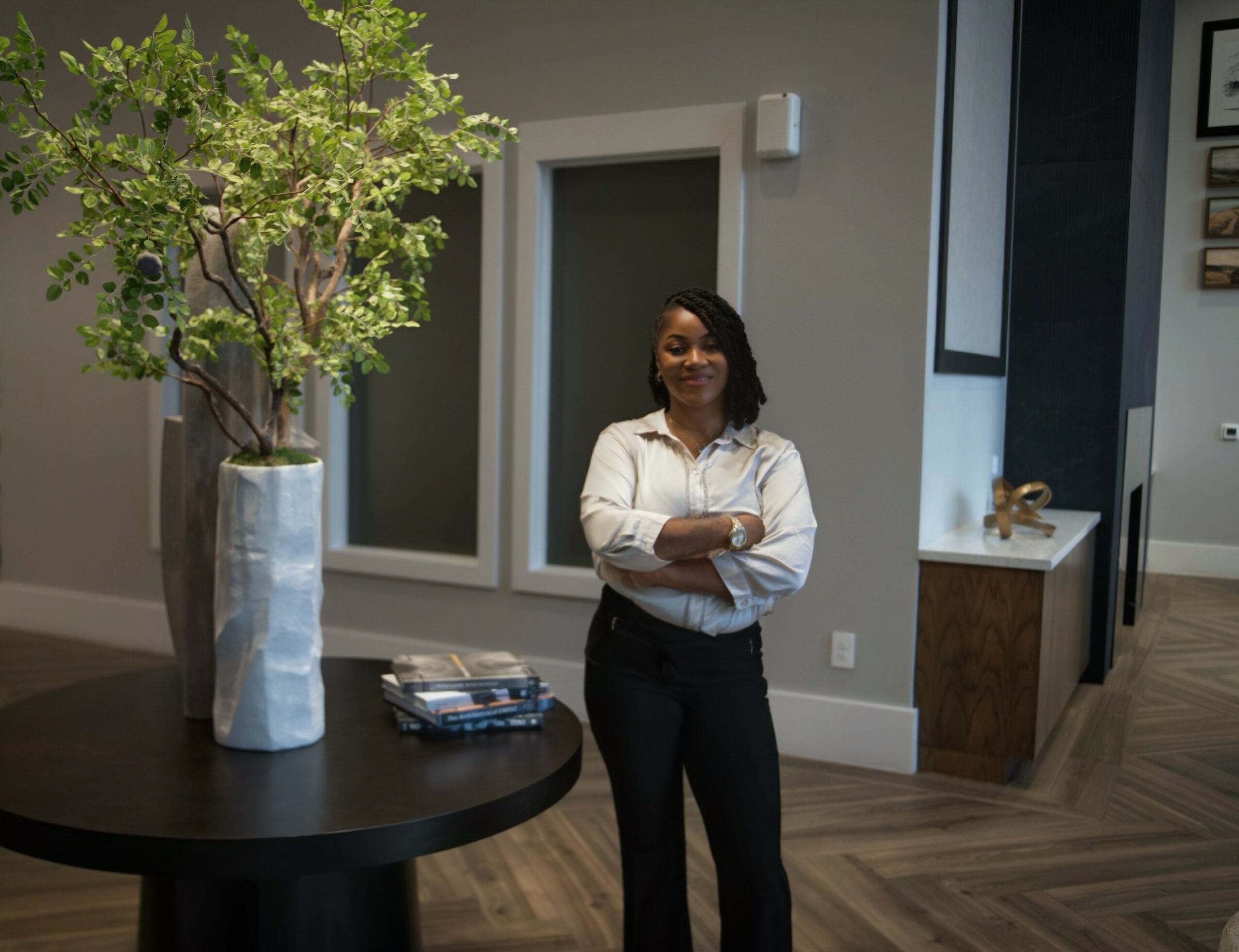 Woman standing with arms crossed in a modern interior with a large plant and artwork, smiling.