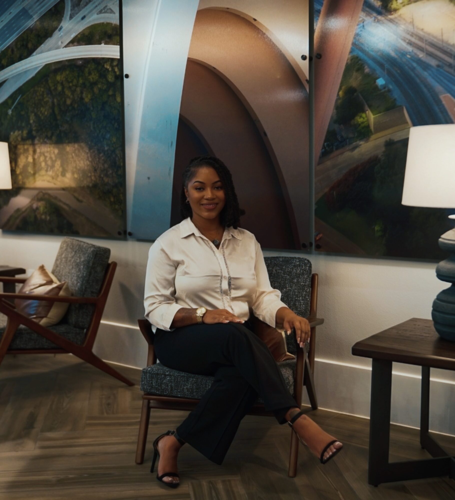Woman sitting on a chair in a modern room with abstract wall art, a side table, and a lamp.
