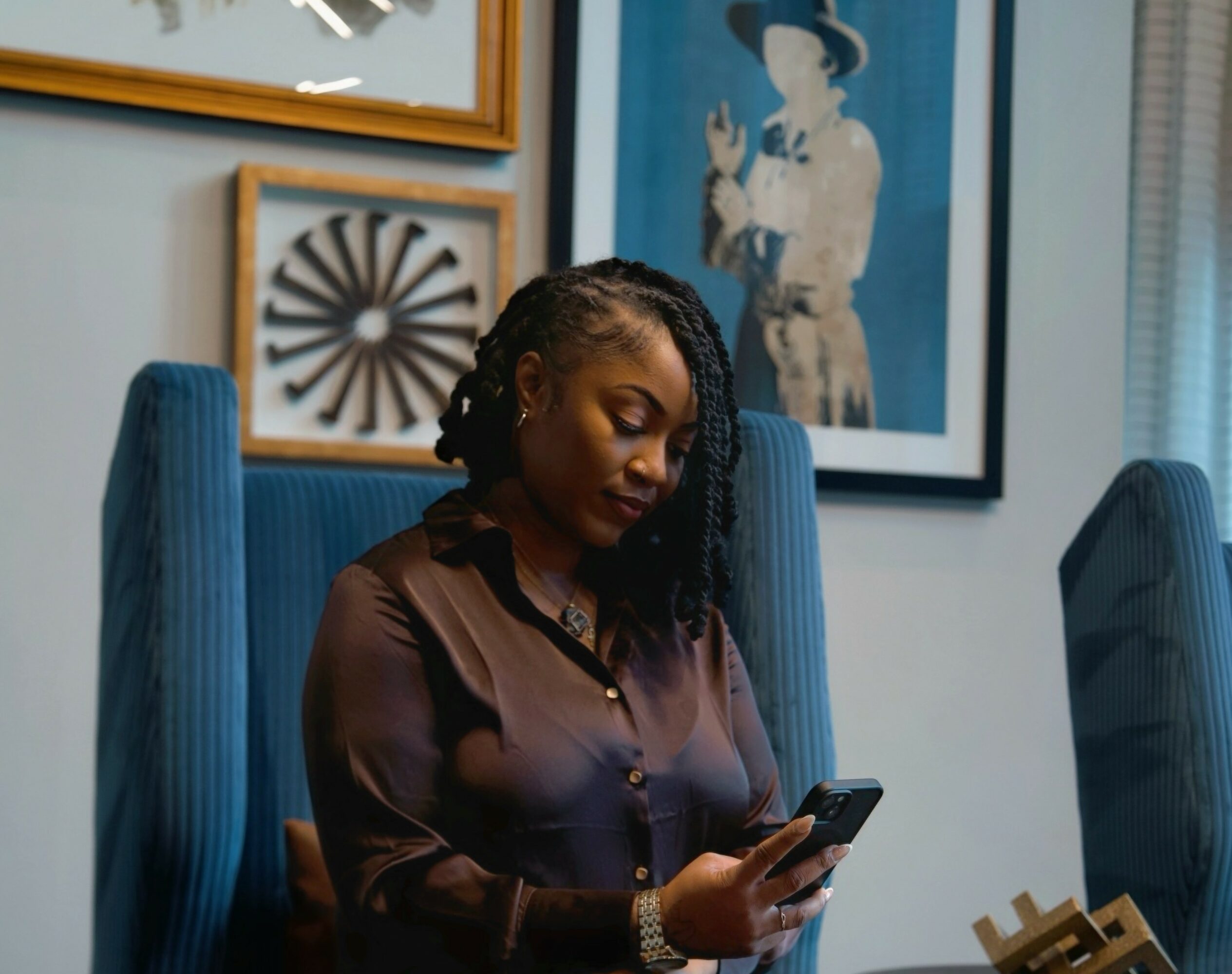 Woman with braided hair looking at her phone, sitting in a room with framed artwork on the wall.