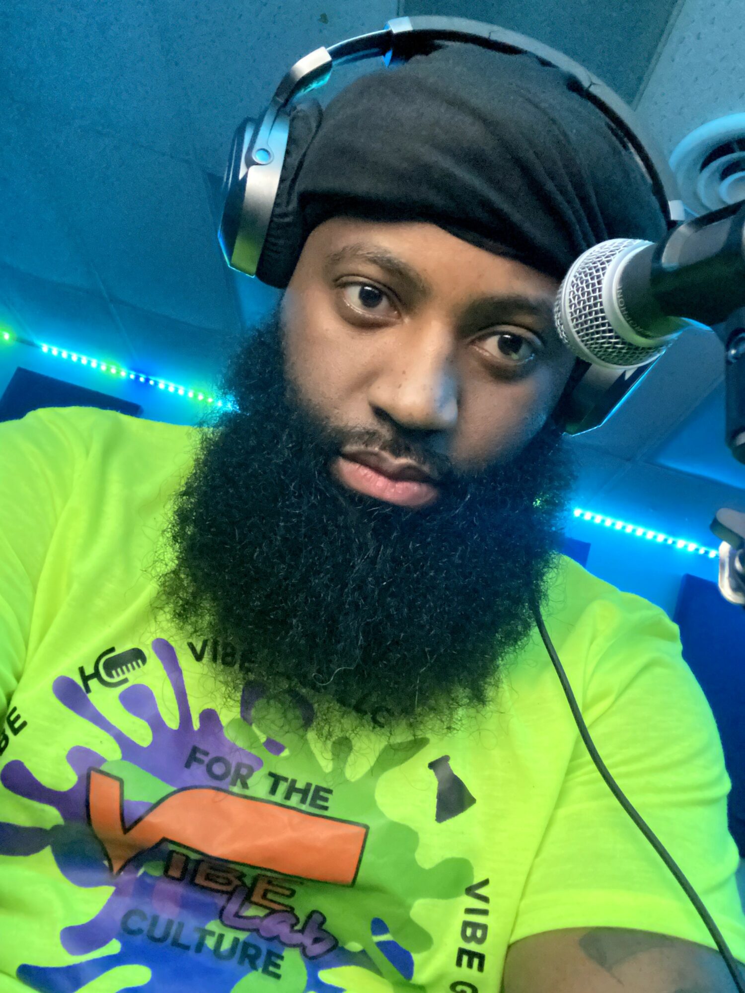 Person with a beard wearing headphones and a black head covering, speaking into a microphone, wearing a bright green shirt with colorful text.
