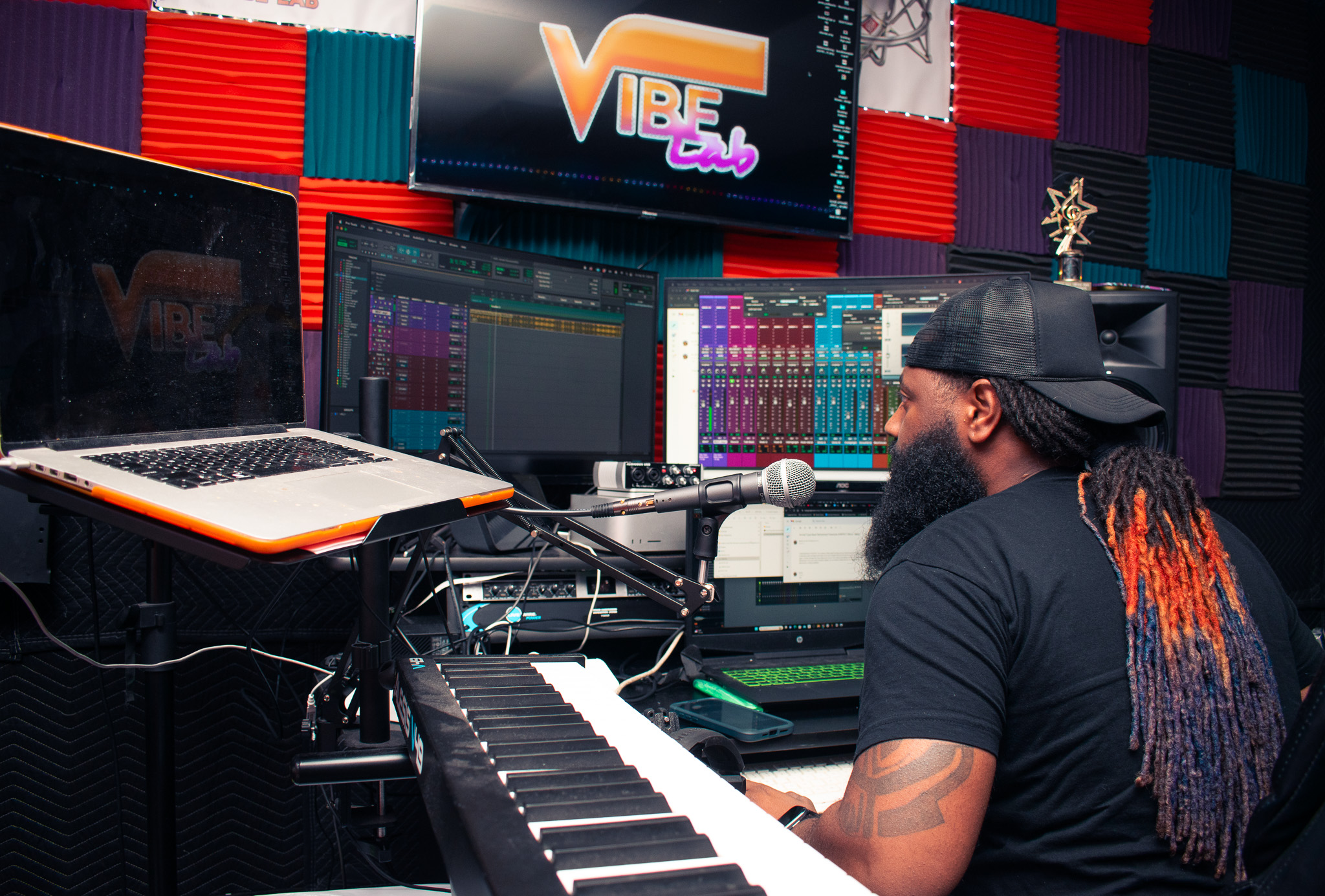Person with long hair and beard wearing a cap, working at a music production setup with keyboard and monitors, in a colorful studio.