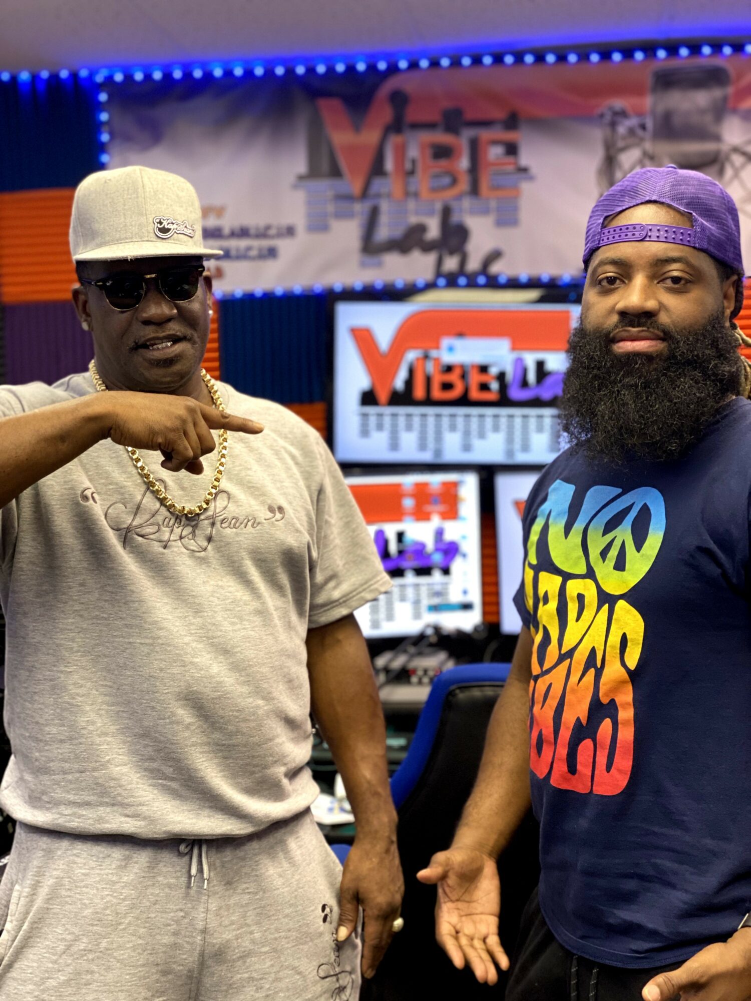 Two men standing in a radio studio with Vibe logo screens in background, one wearing sunglasses and a cap, the other with a beard and colorful shirt.