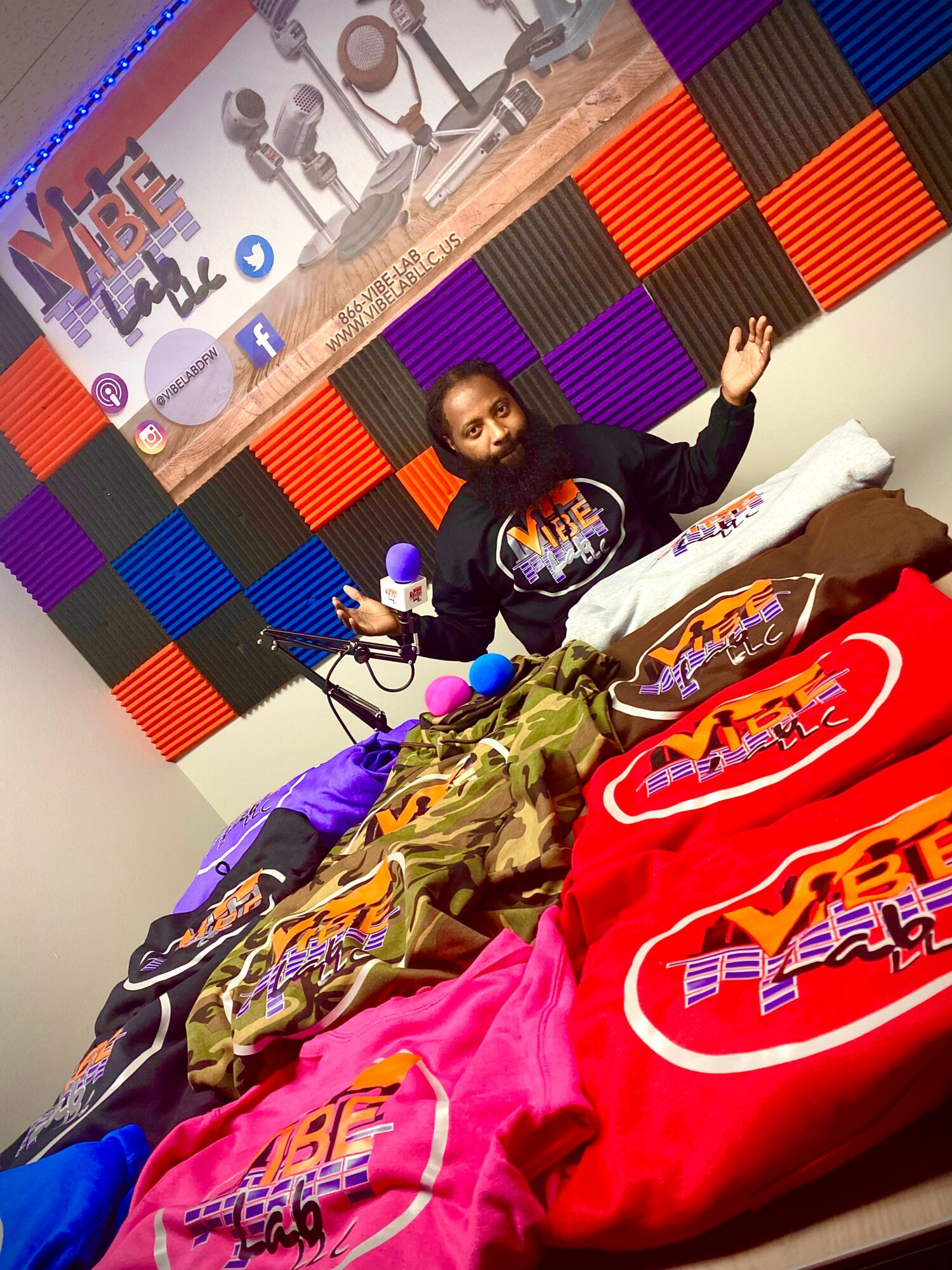 Person sitting on bed with colorful clothing, microphones, and a peace sign gesture, in a room with soundproof panels and a banner.