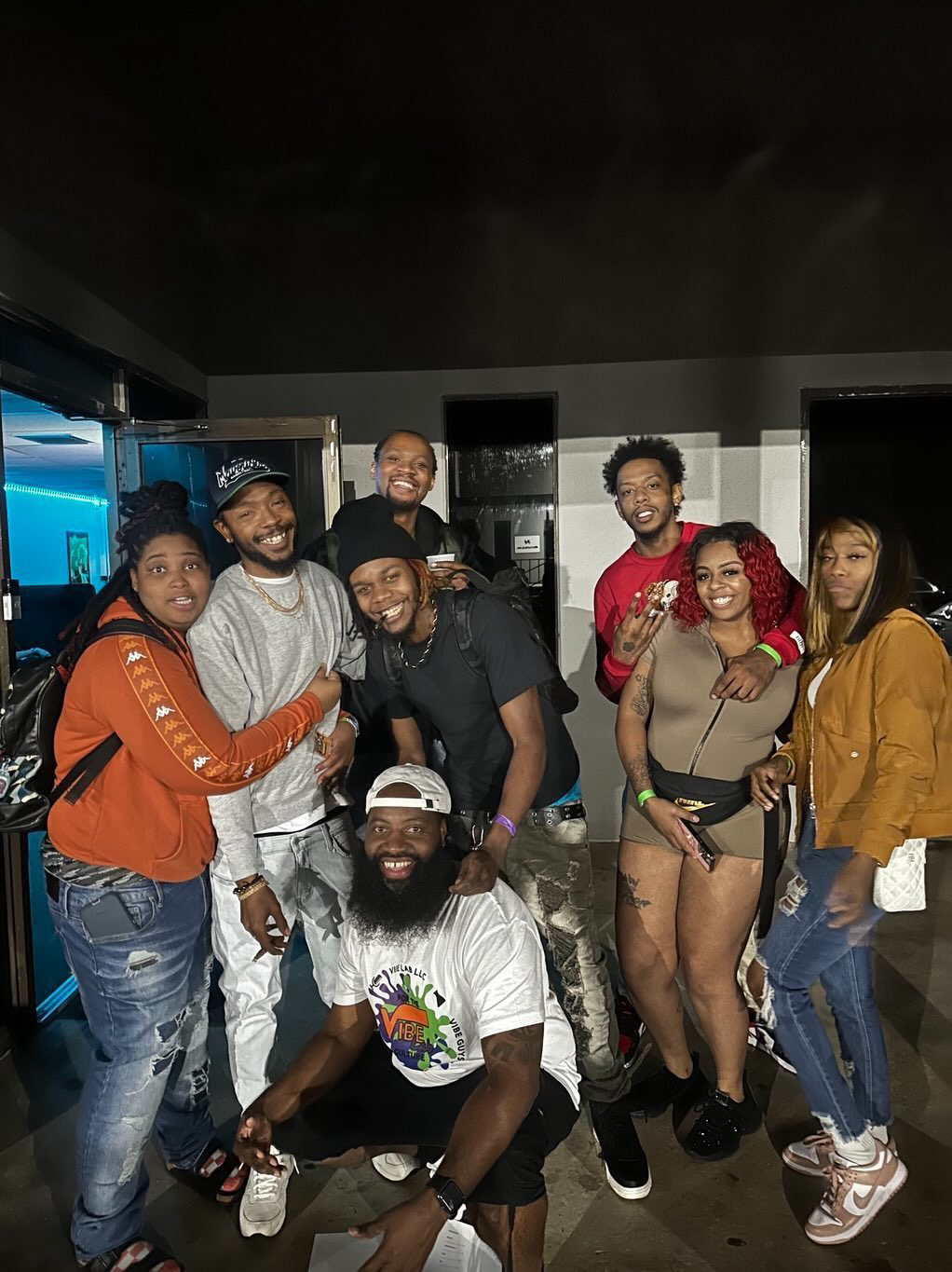 Group of nine people smiling and posing indoors, some with arms around each other, in casual clothing.